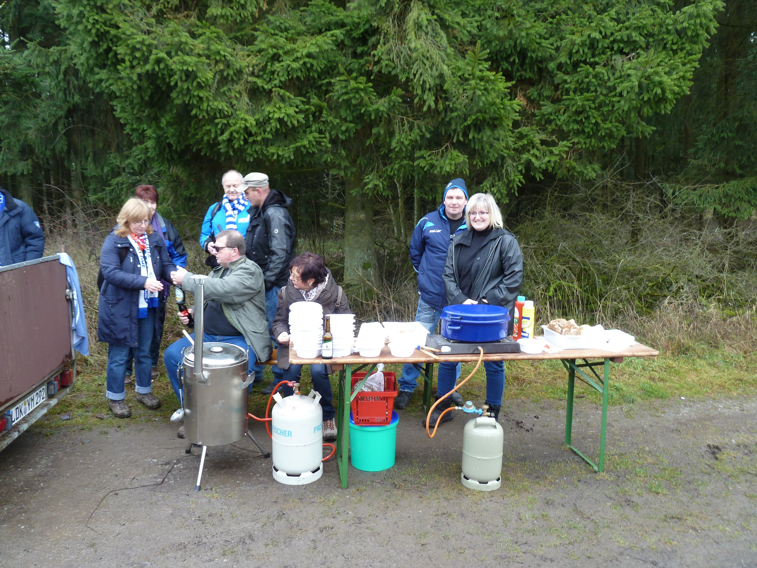 Menschen versammeln sich im Freien bei einem Grill- oder Catering-Stand, umgeben von Bäumen.