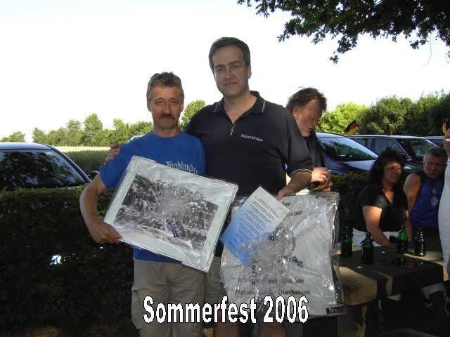 Zwei Männer stehen bei Sommerfest 2006, einen hält er ein gerahmtes Foto und der andere eine Plastiktüte. Im Hintergrund sitzen Menschen an einem Tisch mit Getränken, während es draußen hell und sonnig ist.