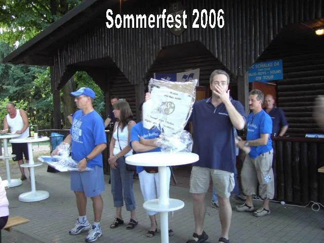 Menschen auf einem Sommerfest 2006 im Freien, einige stehen um einen Stehtisch, einige halten Geschenkpakete, im Hintergrund eine Holzhütte im Schatten.