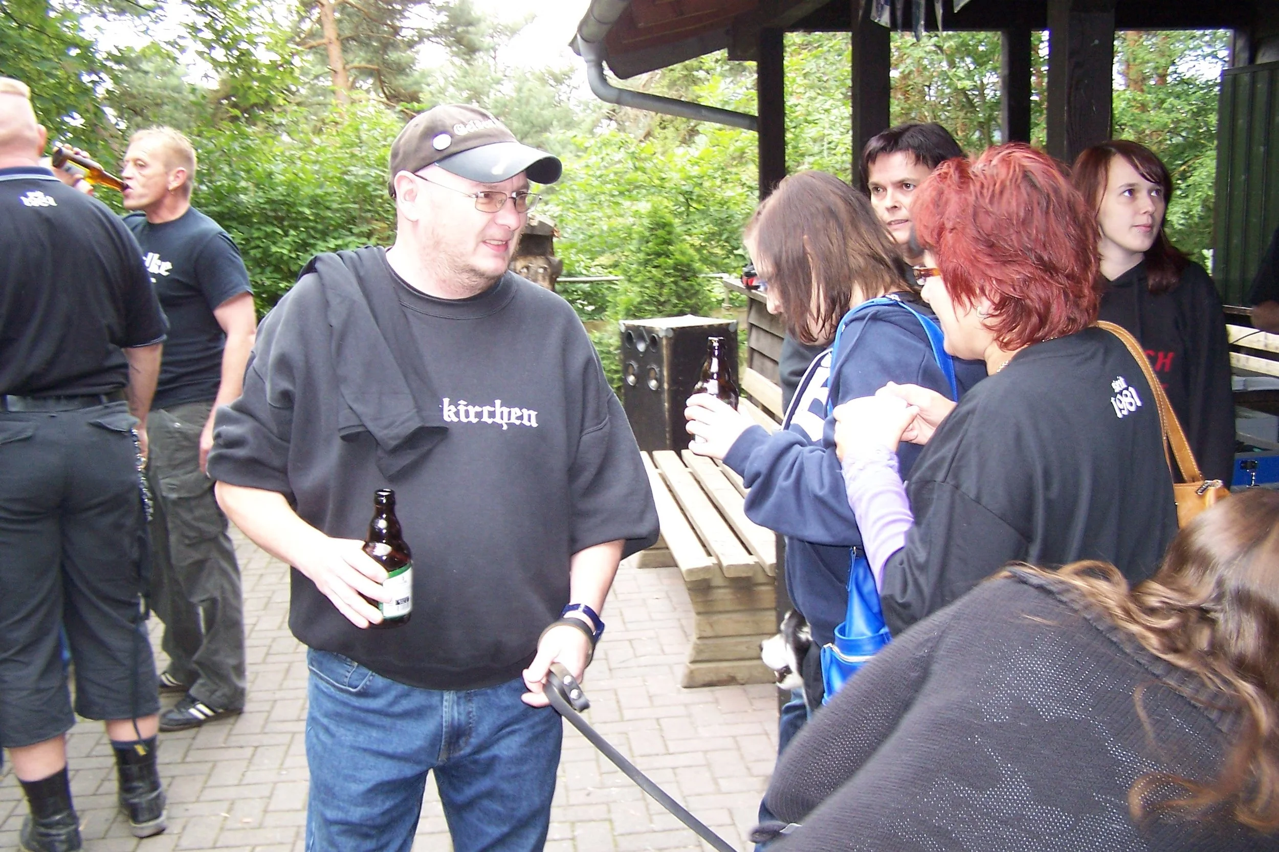 Gruppe von Menschen, die sich bei einem Outdoor-Treffen unterhalten, einige halten Getränke, im Hintergrund ist eine grüne, bewaldete Umgebung.
