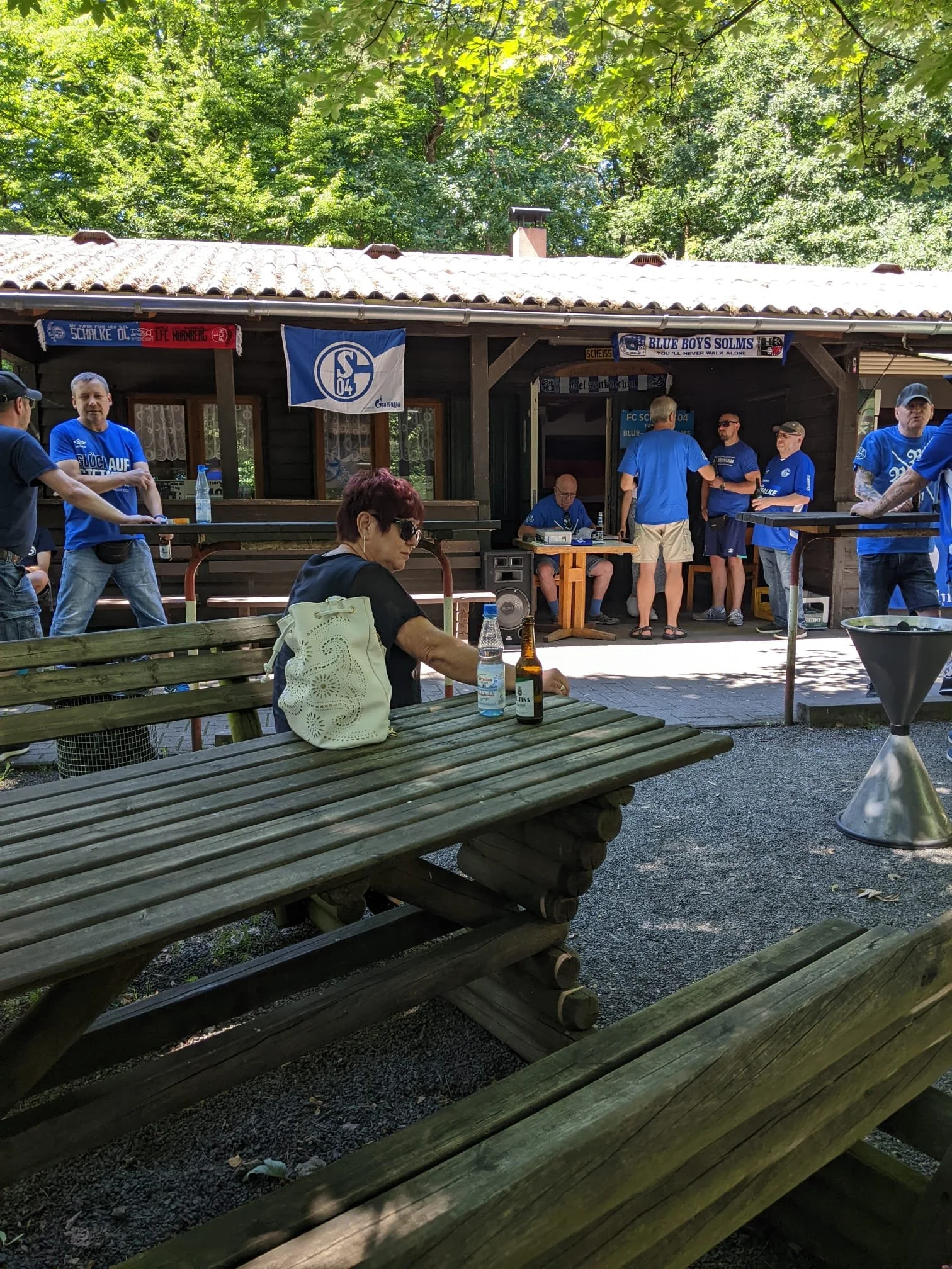 Menschen in blauen T-Shirts sitzen und stehen vor einer kleinen Hütte, umgeben von grünen Bäumen, bei einer Veranstaltung, mit einigen Tischen und Bänken im Freien.