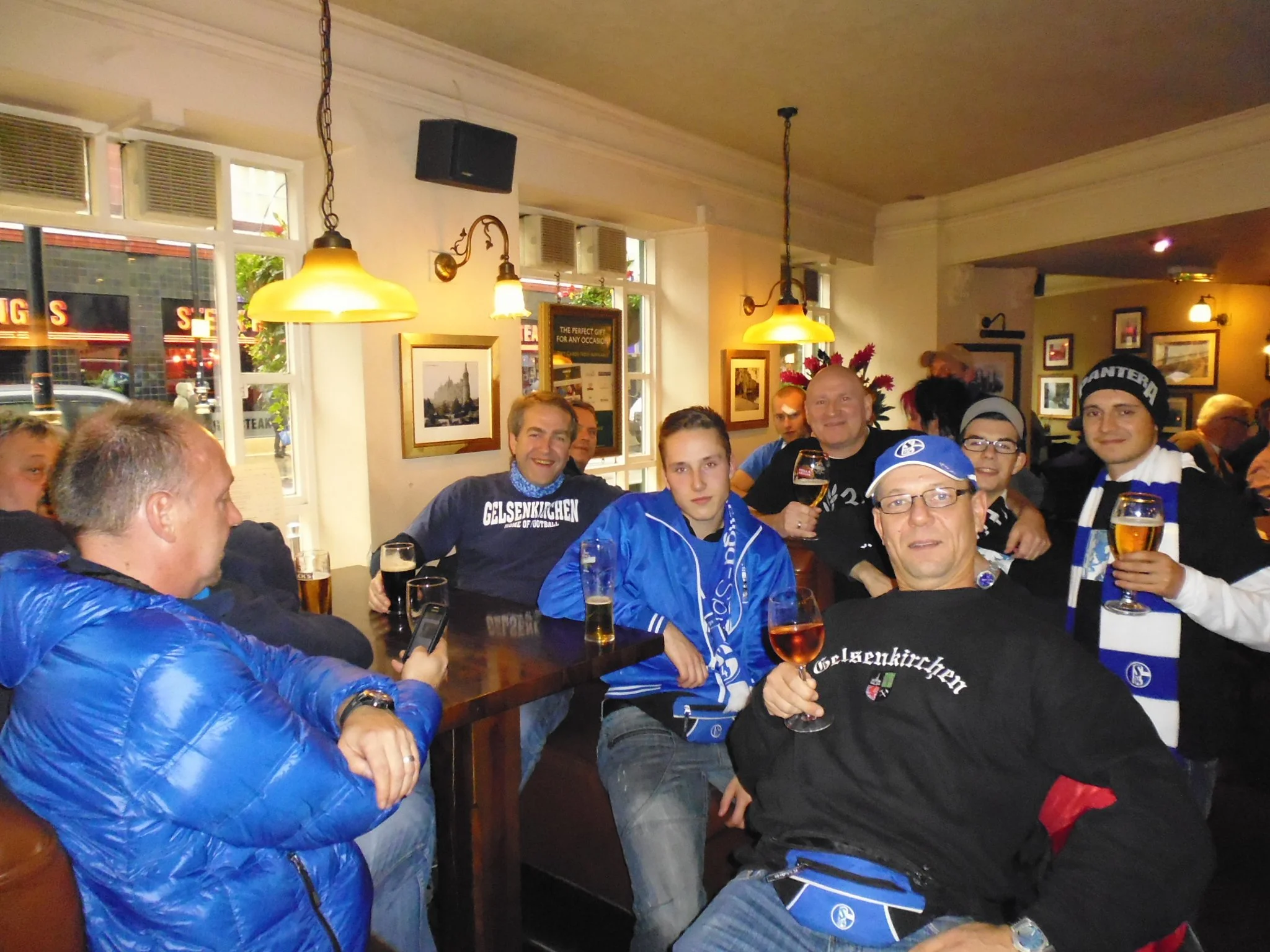 Mehrere Personen in einer Bar oder einem Restaurant, auf der rechten Seite eine Gruppe von Menschen, die Getränke und Bier trinken, einige tragen Schal und Mützen, im Hintergrund fenster, Bilder an der Wand, und Deckenleuchten.