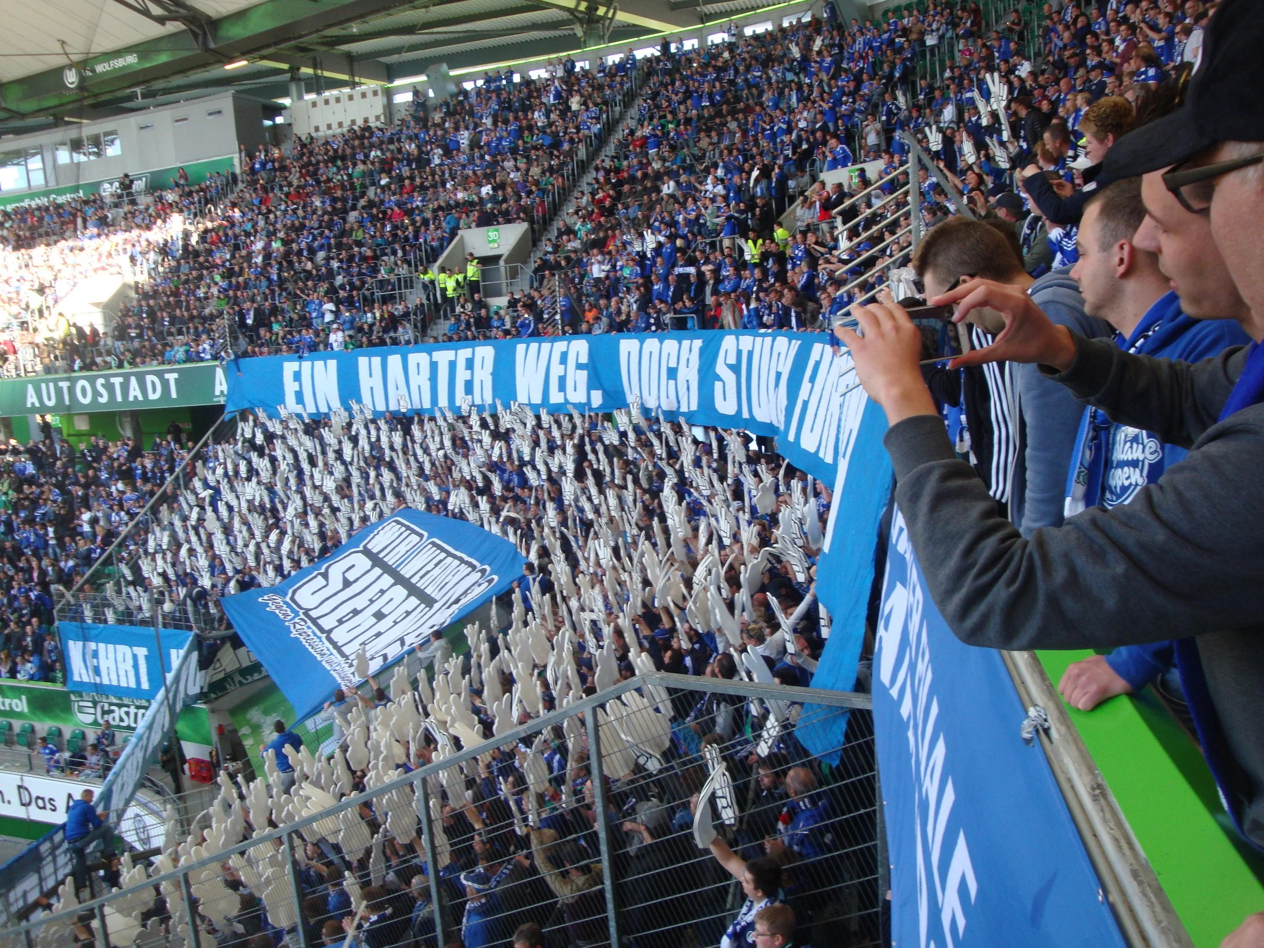 Fußballfans in einem Stadion, viele tragen blaue Kleidung und halten Schwenkfahnen, während sie das Spiel beobachten.