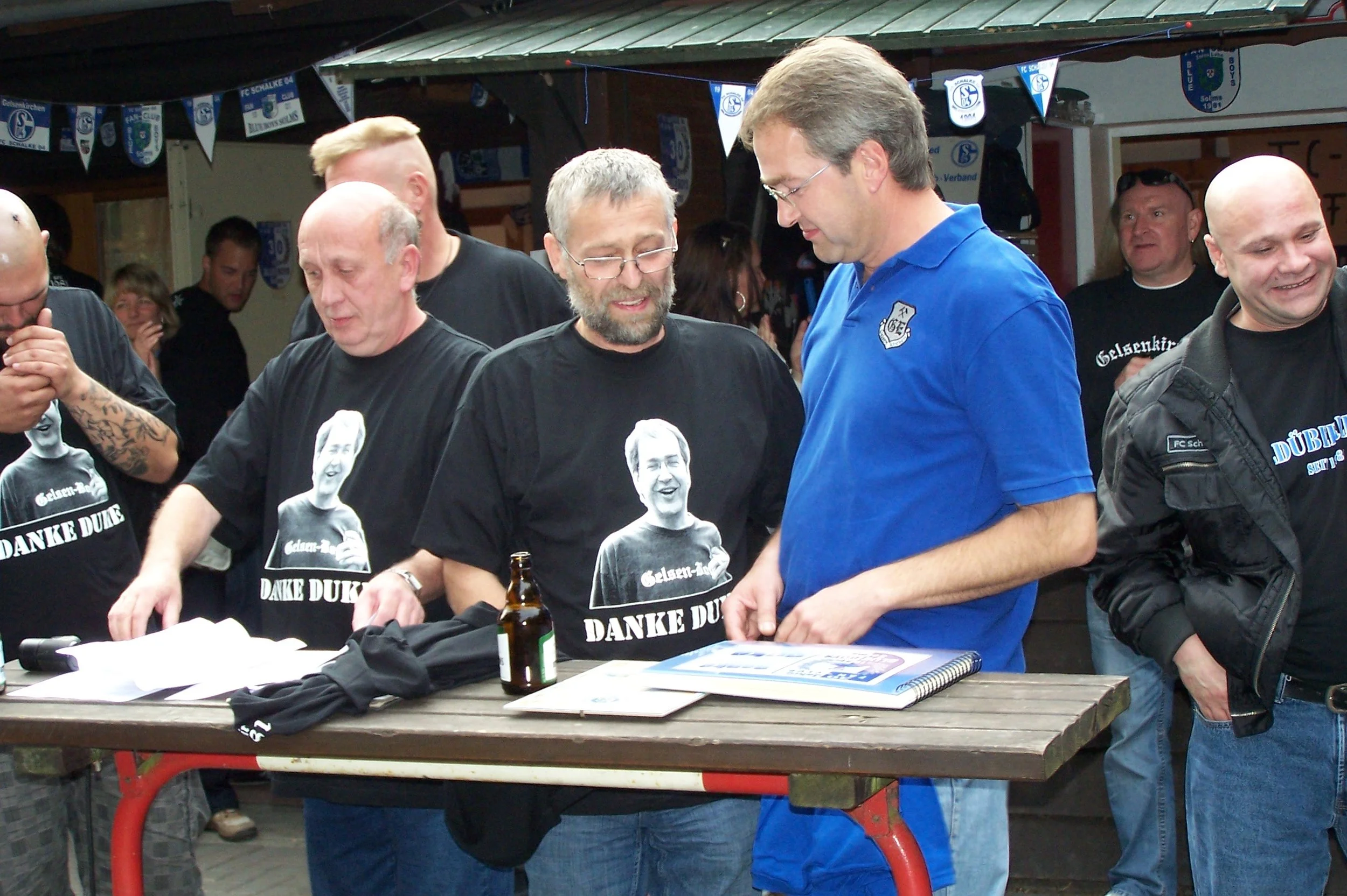 Mehrere Männer stehen um eine Theke im Freien, einige tragen schwarze T-Shirts mit weißen Druckmotiven und Text, während einer eine blaue Polo-Hemd trägt. Im Hintergrund hängen blaue und weiße Fahnen und Banner mit Vereinslogos. Es sieht nach einer g