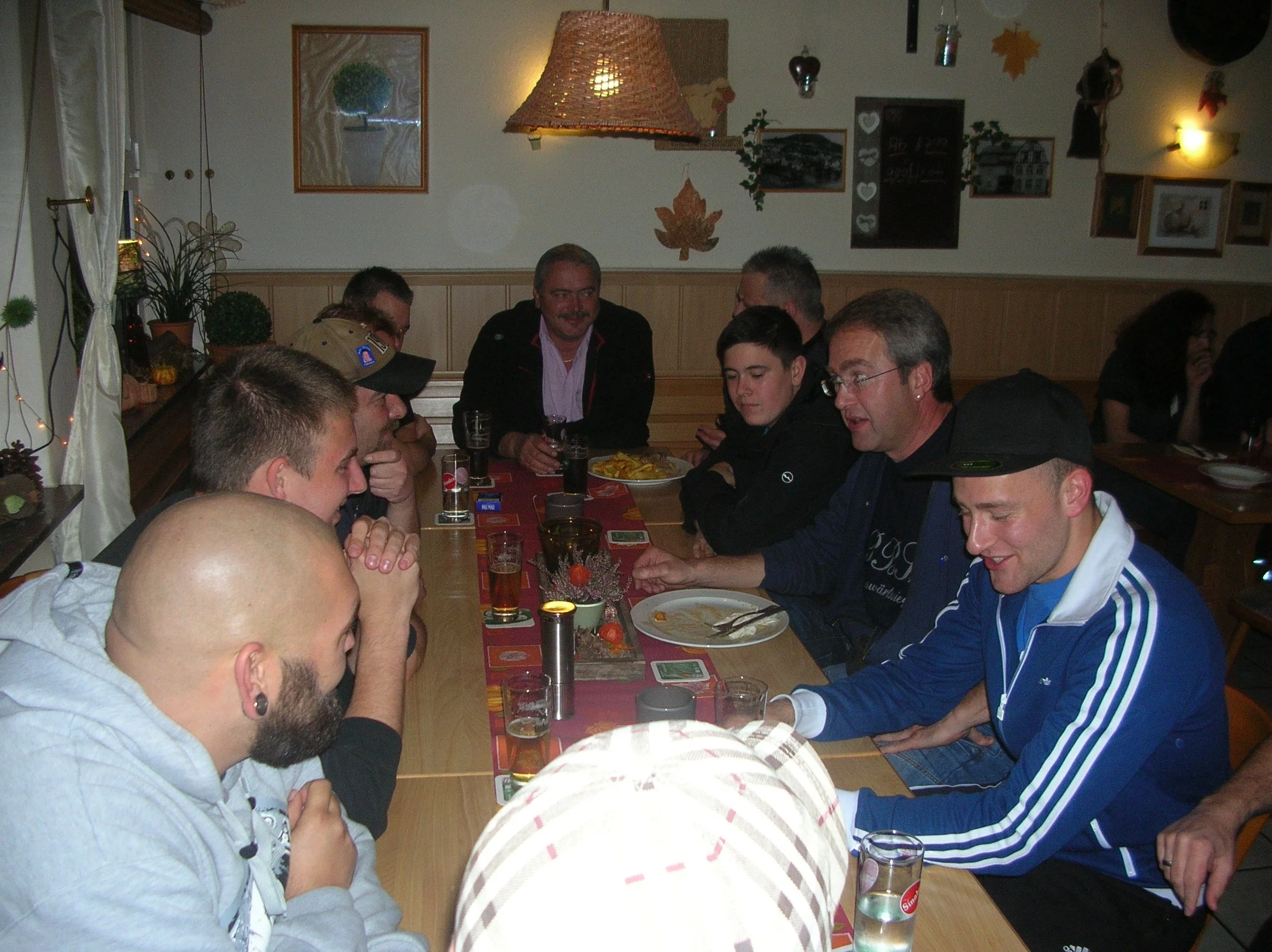 Mehrere Menschen sitzen an einem langen Tisch in einem gemütlichen Raum, einige essen, andere unterhalten sich. Es sieht nach einer kleinen Feier oder einem Treffen aus, mit hellen Lichtern und Pflanzen an der Wand.