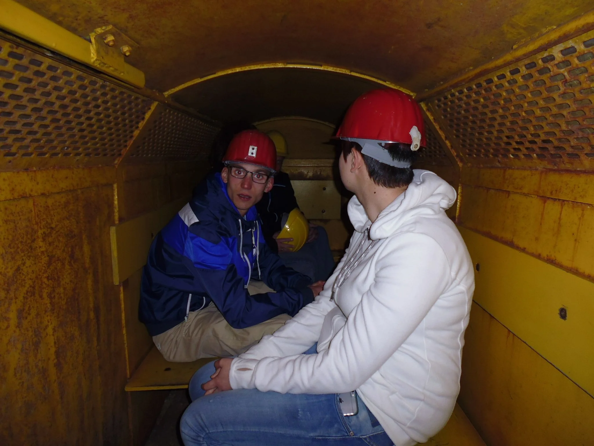 Zwei Personen mit roten Helmen sitzen in einem gelb gestrichenen Tunnel oder Korridor aus Metall, die eine Person schaut direkt in die Kamera, während die andere Person nach hinten schaut.