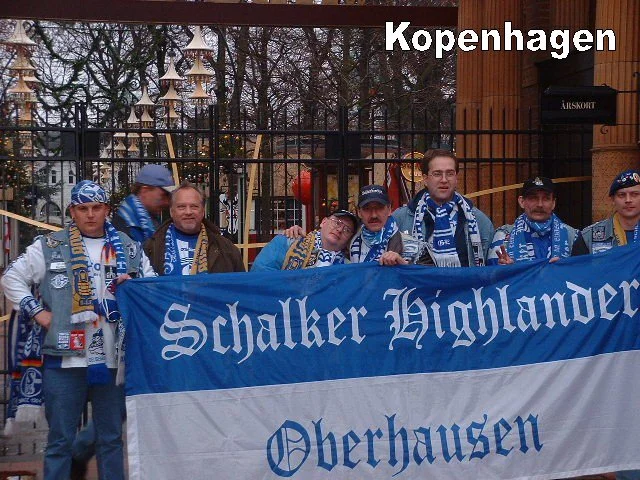 Eine Gruppe von sieben Menschen, die Schalke-Highlander- und Oberhausen-Fans in blauen und weißen Schals und Kleidung tragen, hält ein großes blau-weißes Banner mit der Aufschrift "Schalker Highlander Oberhausen" vor einem Zaun mit Weihnachtsdeko im 