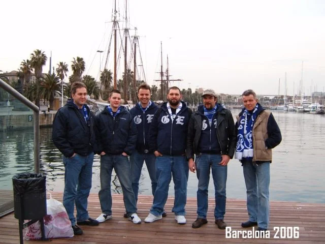 Sechs Männer stehen auf einem Dock am Wasser, im Hintergrund sind Boote und Palmen am Hafen in Barcelona, 2006.