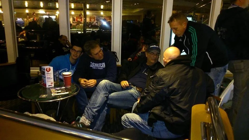 Fünf Männer sitzen und stehen in einer Wartehalle oder einem Cafè, an einem runden Tisch mit einigen Zeitschriften, Stiften und einem Pappbecher, während sie sich unterhalten.