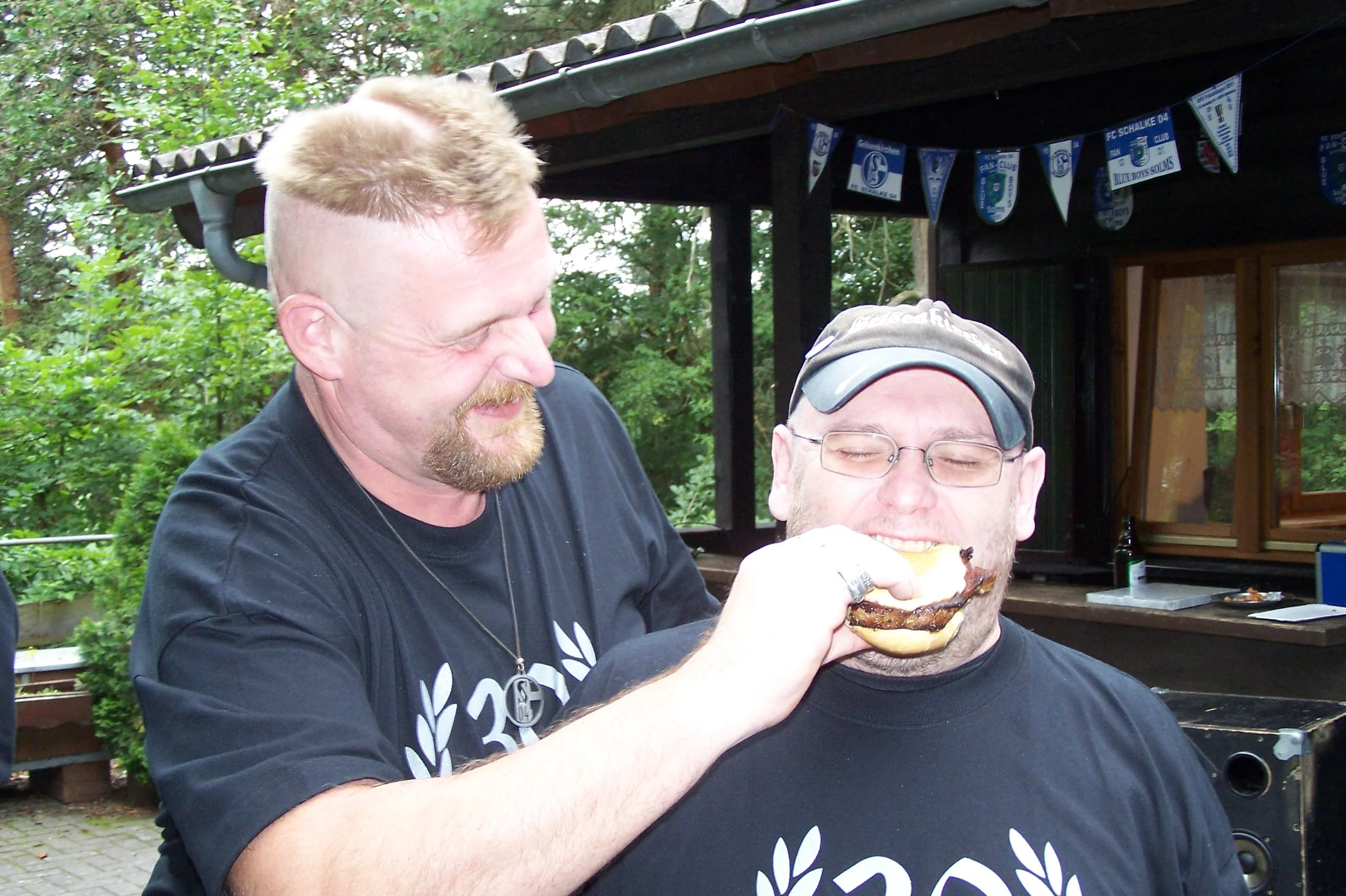 Zwei Männer essen beim Grillen im Freien, mit Bäumen und einem kleinen Häuschen im Hintergrund.