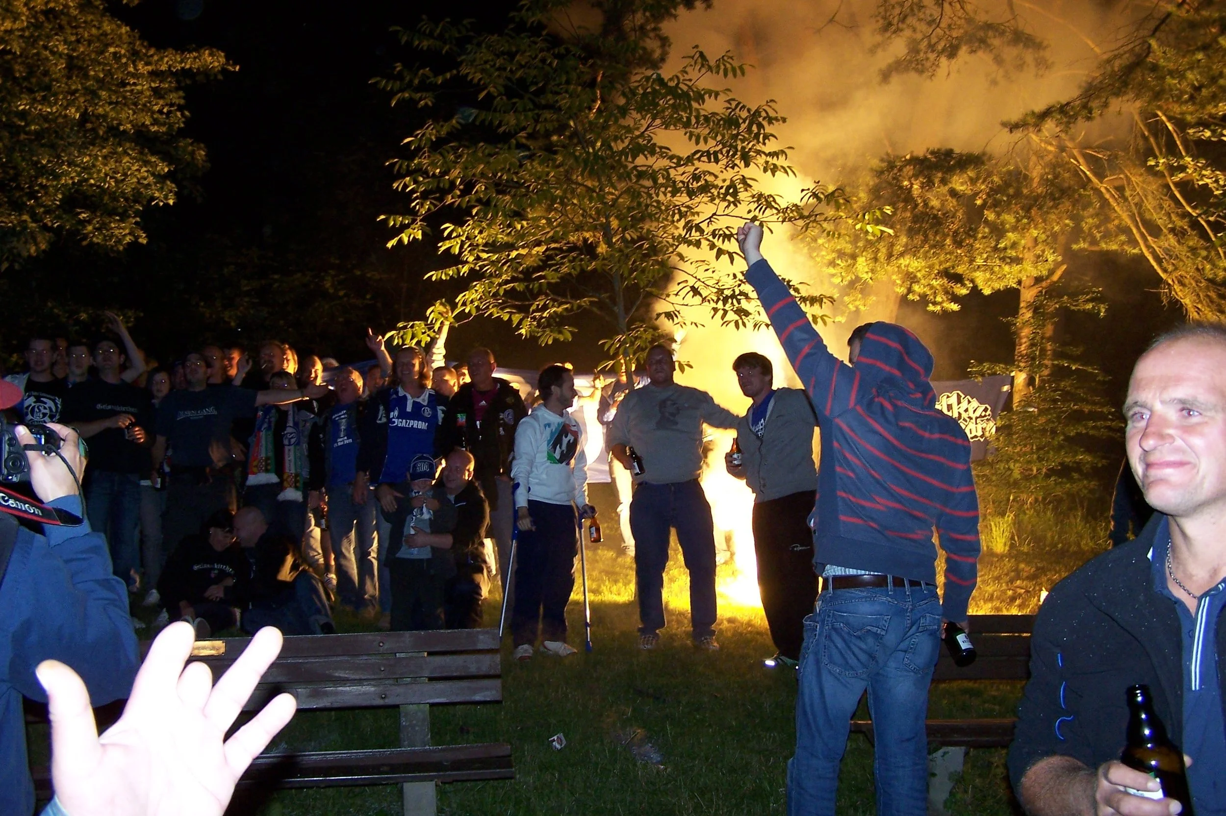 Menschen versammeln sich nachts im Freien vor einem brennenden Feuer, einige feiern und trinken Bier, umgeben von Bäumen.