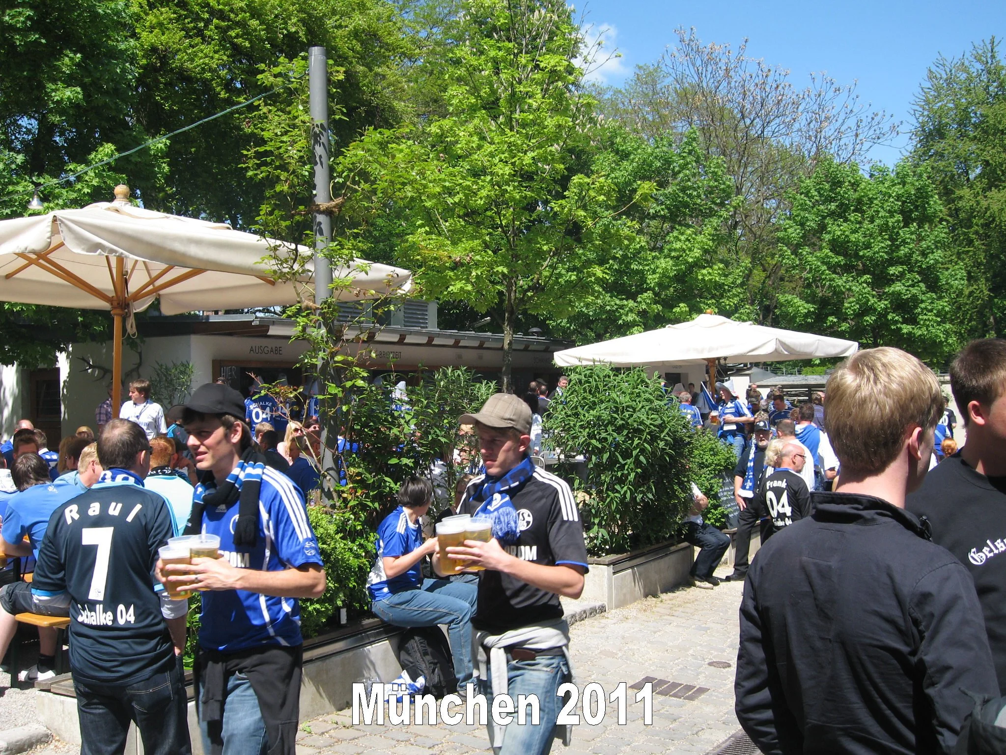 Menschen in blauen Trikots feiern im Freien vor einem Baum mit grünen Blättern, einige halten Biergläser. Im Hintergrund sind grüne Bäume, ein Gebäude und Sonnenschirme. Text auf dem Bild: 'München 2014'.