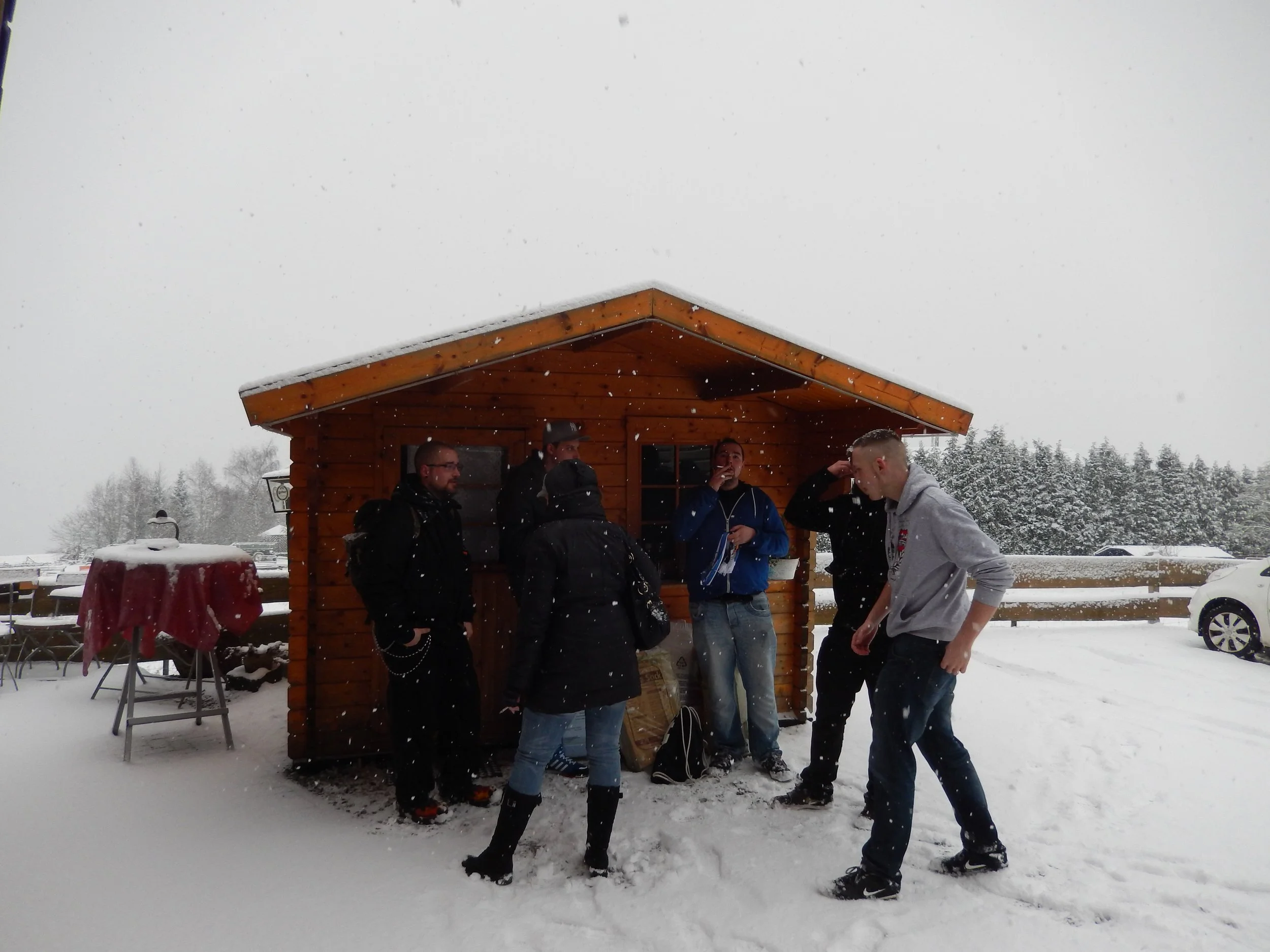 Gruppe von Menschen, die vor einem kleinen Holzhaus im Schnee stehen, bei Schnee und über Wolken.