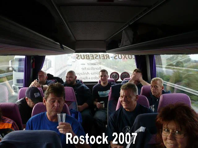 Gruppen von Menschen sitzen in einem Bus, einige trinken, andere sprechen. Text auf der Rückscheibe lautet 'Rostock 2007'.