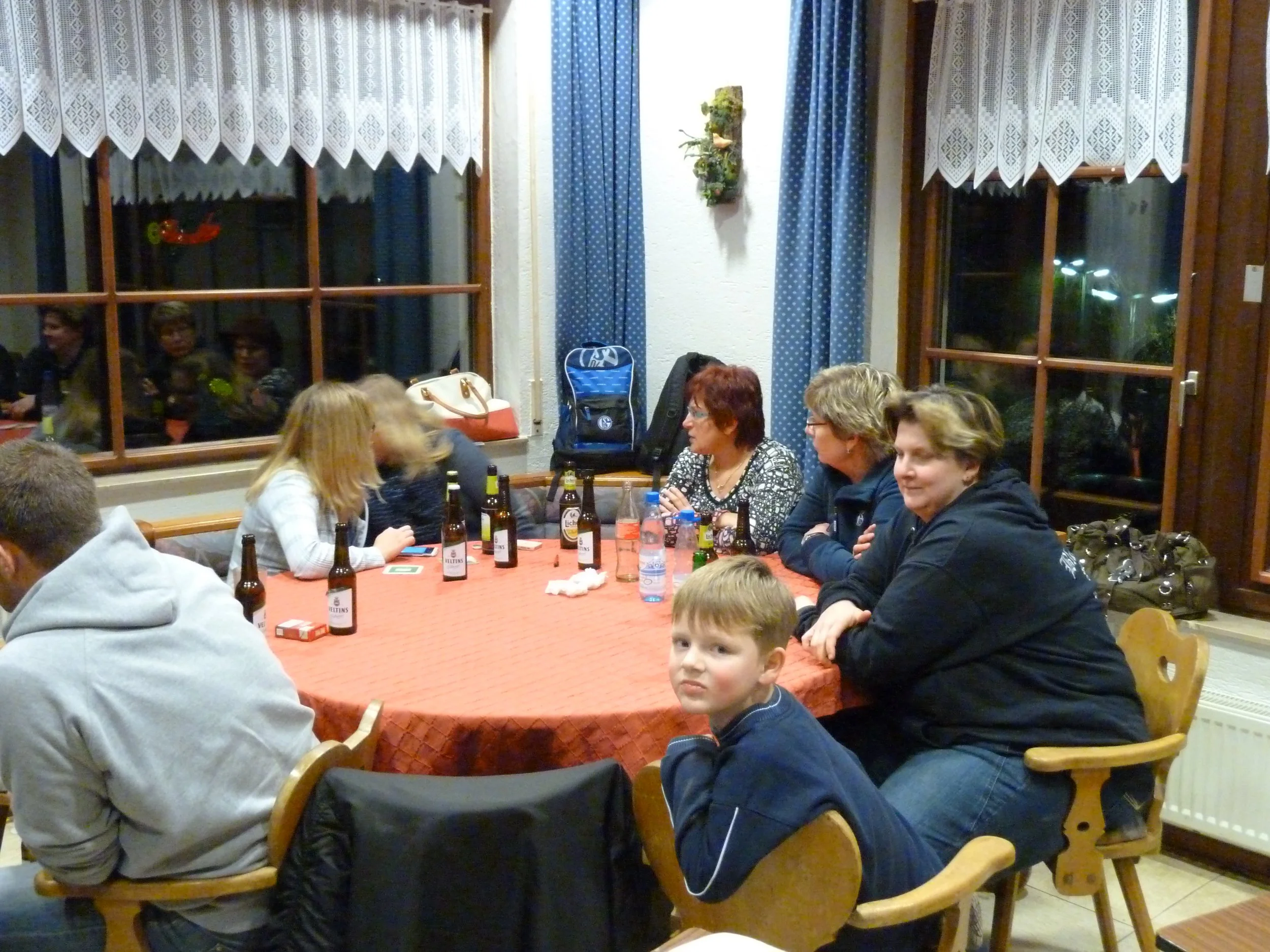 Menschen sitzen um einen runden Tisch mit roten Tischtuch und trinken Bier. Es ist eine festliche Umgebung mit mehreren Frauen und einem Kind im Vordergrund.
