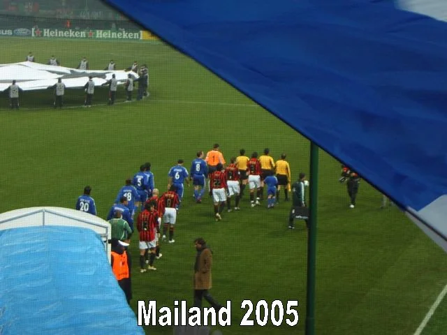 Fußballspieler laufen auf dem Spielfeld bei einem Spiel in Mailand 2005, mit Zuschauern und Zelten im Hintergrund.
