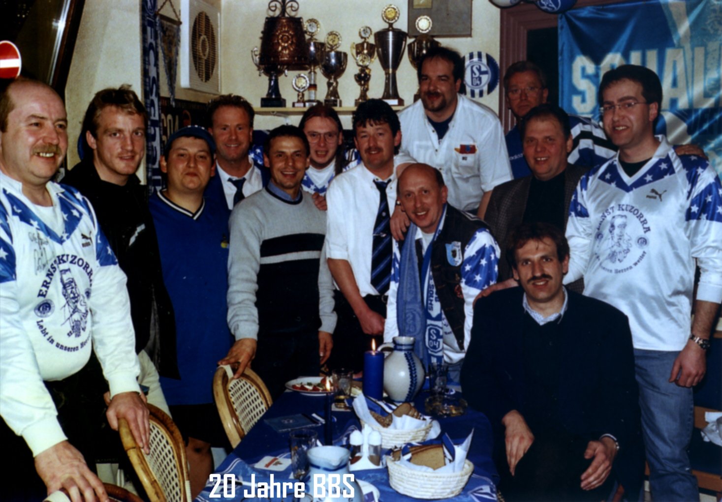 Gruppe von Fußballfans in Vereinshaus, alle tragen Fan- oder Vereinskleidung, im Vordergrund ein Mann sitzt an einem festlich gedeckten Tisch, im Hintergrund Trophäen und Vereinsbanner, auf dem Bild steht '20 Jahre BBS'.