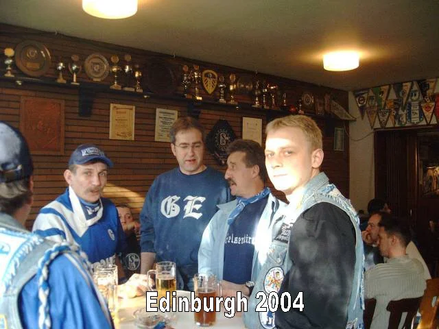 Menschen in einem Pub, einige tragen Vereinskappen und -shirts, im Hintergrund Wanddeko mit Pokalen und Stickern, Text 'Edinburgh 2004'