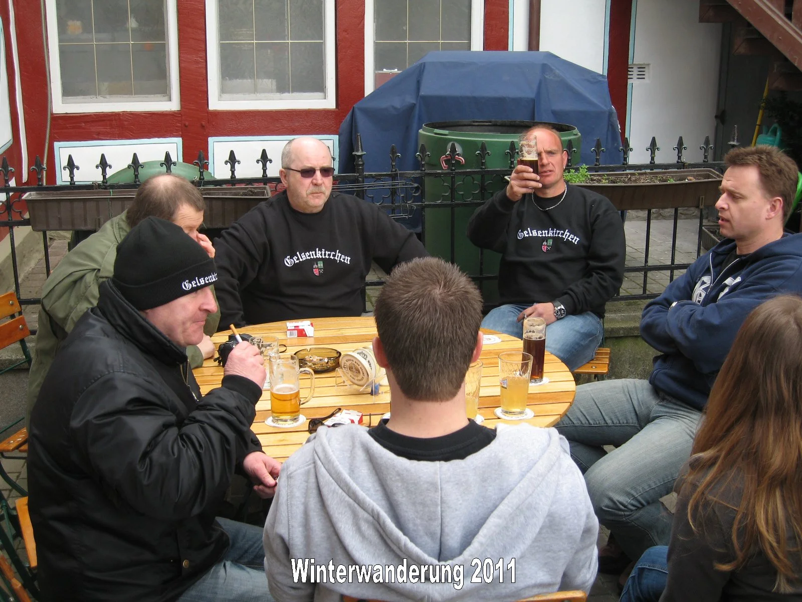 Eine Gruppe von Menschen sitzt im Freien an einem runden Holztisch und trinkt Bier, einige tragen schwarze Sweatshirts mit dem Schriftzug 'Gelsenkirchener'.