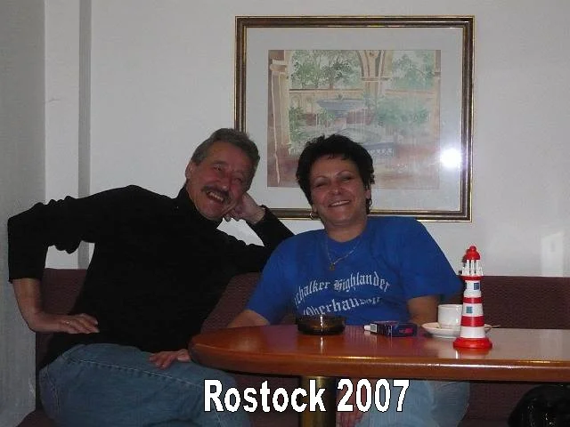 Zwei lachende Menschen, ein Mann und eine Frau, sitzen an einem Tisch in Rostock im Jahr 2007, mit einer Wasserstanderkennung und einem Fotoshop-Logo im Bild.