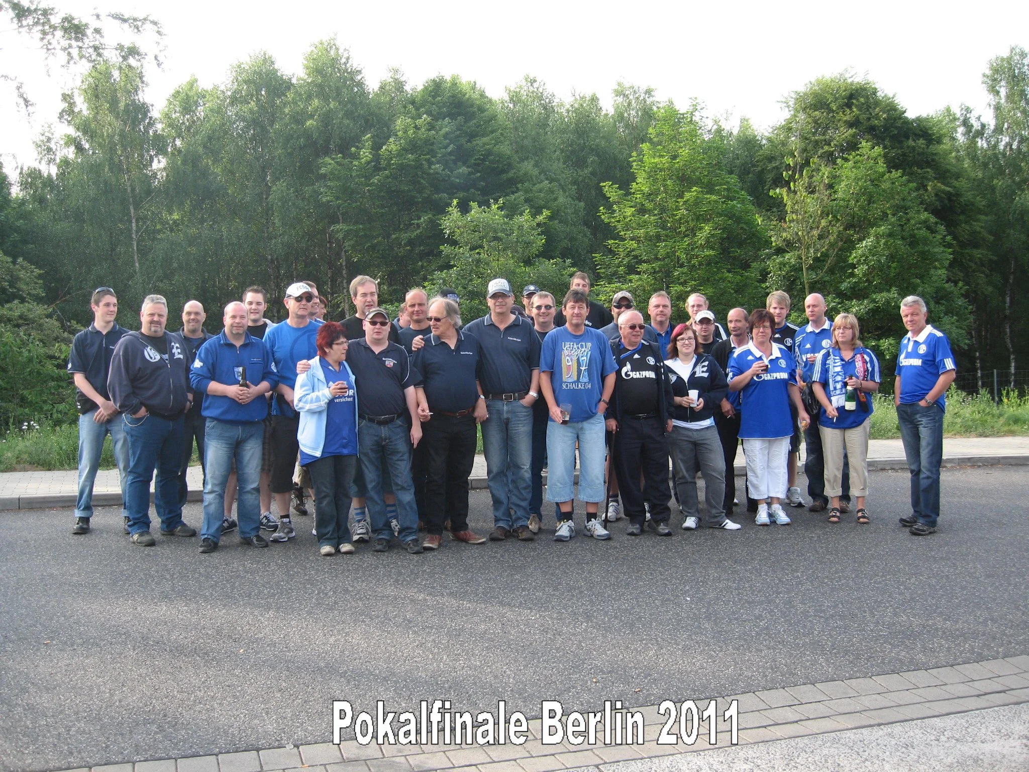 Gruppe von Menschen in blauen und schwarzen Kleidern vor einer grünen Baumkulisse, einige halten Getränke, vermutlich beim Pokalfinale Berlin 2011.