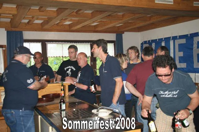 Menschen beim Sommerfest 2008, einige halten Getränke, im Hintergrund ein Banner und ein Fenster mit Blick nach draußen.
