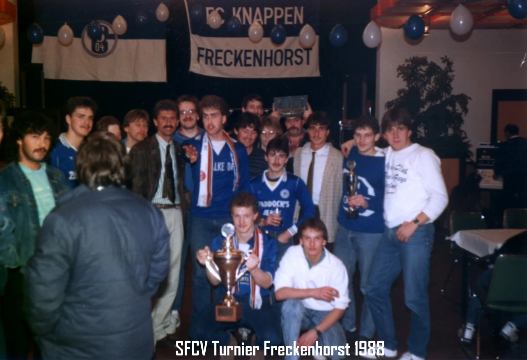 Gruppen von jungen Männern feiern einen Sieg bei einem Turnier 1988, in der Mitte halten sie einen Pokal, im Hintergrund ein Banner mit der Aufschrift 'FC Knappen Freckenhorst'.
