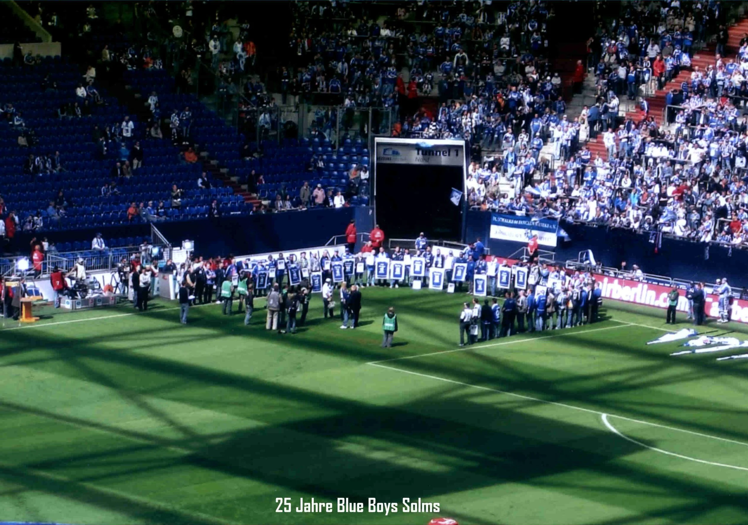 Fußballstadion mit Zuschauern auf den Tribünen, einige stehen auf dem Spielfeld, Menschen in blauen Trikots, tribünenweiten Banner, im unteren Bereich Text: '25 Jahre Blue Boys Solms'.