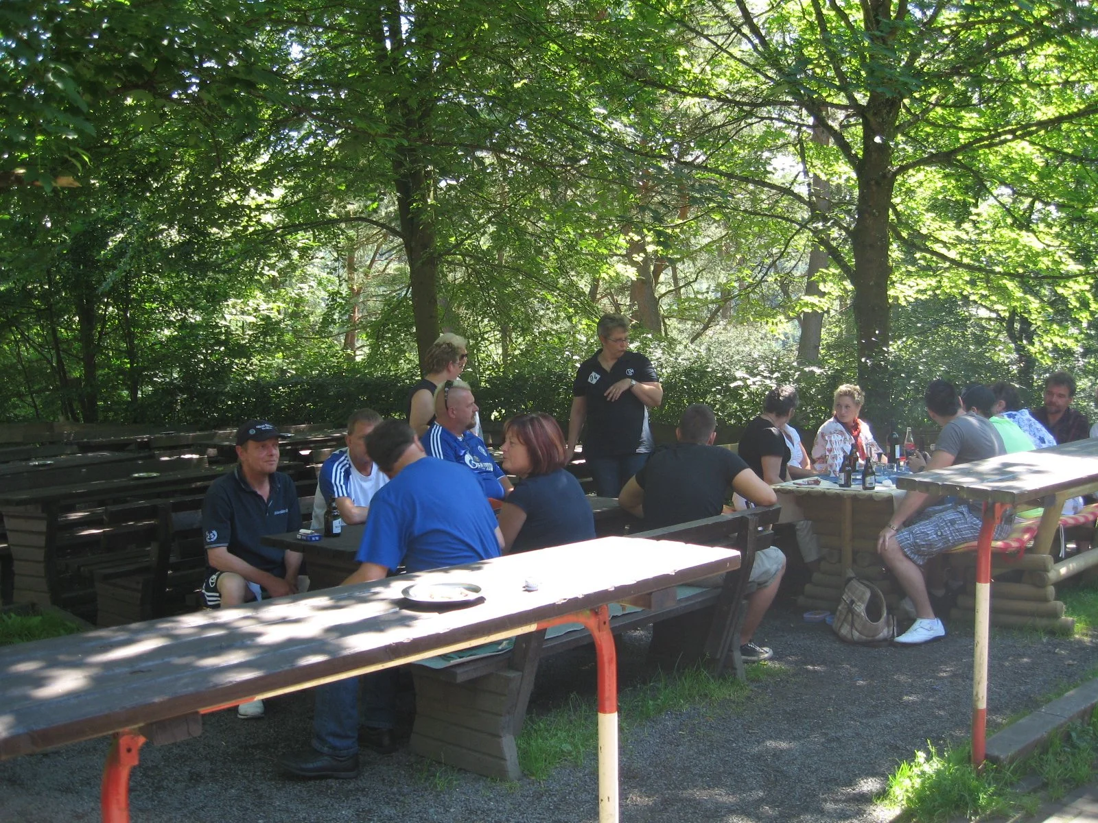 Menschen sitzen an Tischen im Freien unter Bäumen bei einem Treffen oder einer Feier.