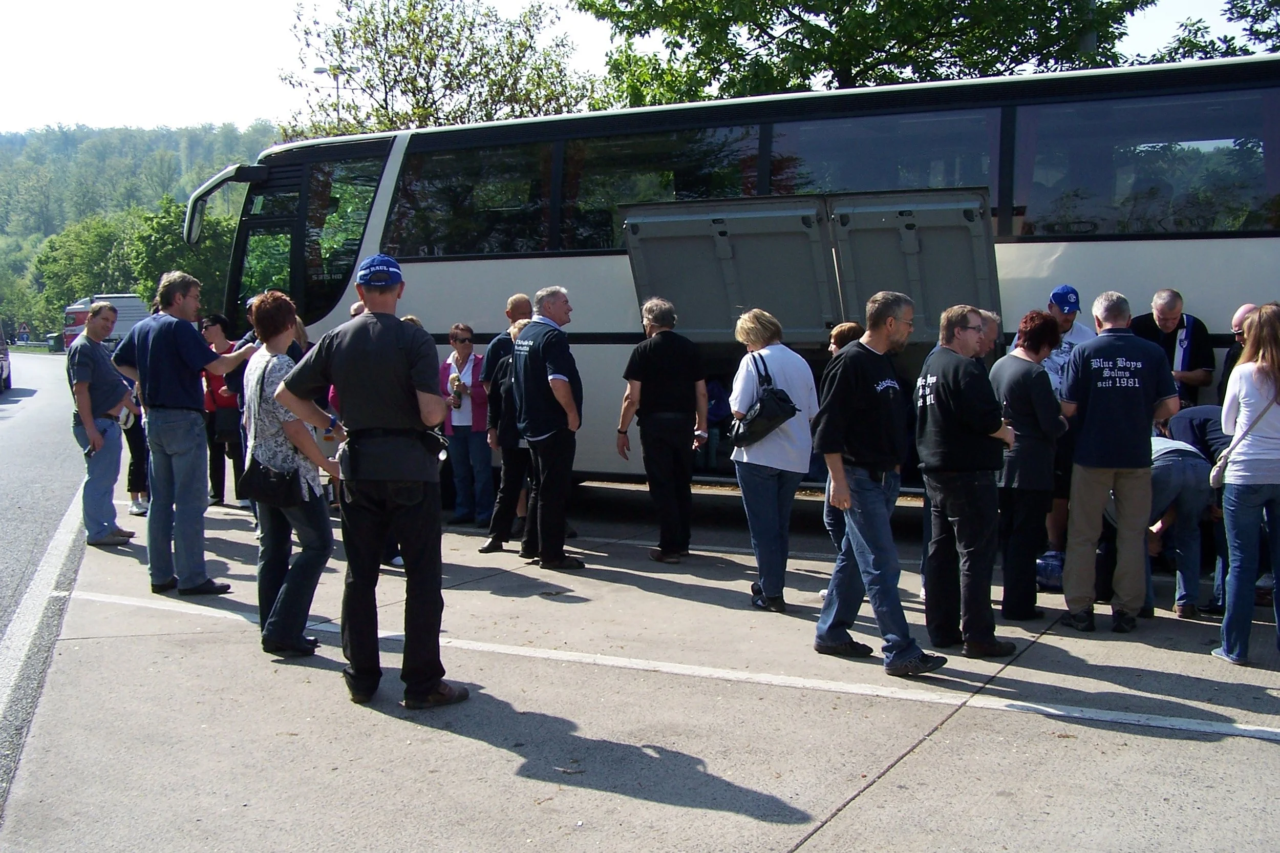 Menschen stehen in einer Gruppe in der Nähe eines großen Busses im Freien, mit Bäumen und einem Himmel im Hintergrund.
