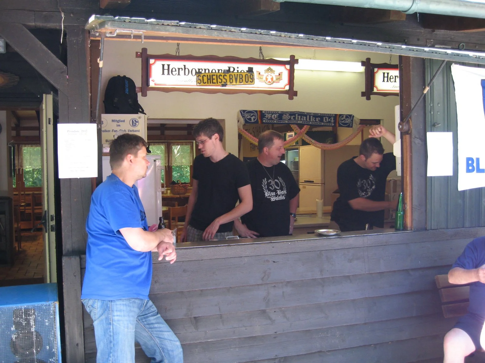 Blick in einen Holzbar mit mehreren Männern, die vor einem offenen Fenster stehen, darin Getränke und einen Kühlschrank. Das Schild über dem Fenster liest 'Herzöppen Bier' und hat einen irrtümlichen Schreibfehler. Im Hintergrund sind weitere Dekorati
