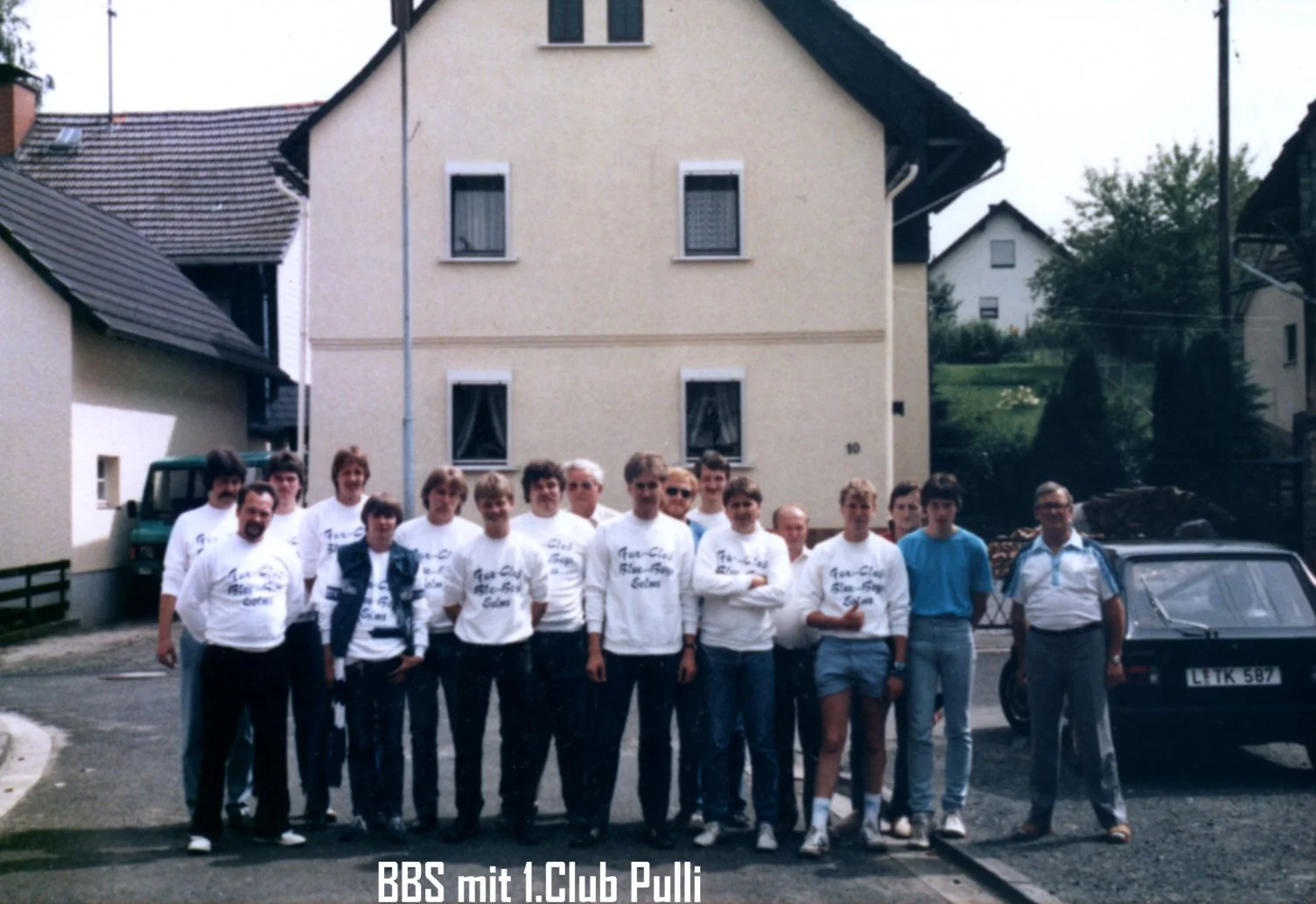 Gruppenfoto von mehreren Männern und Frauen vor einem Haus. Einige tragen T-Shirts mit Aufdruck, wahrscheinlich im Zusammenhang mit einem Club oder Verein. Es scheint sich um eine Familien- oder Vereinszusammenkunft zu handeln.