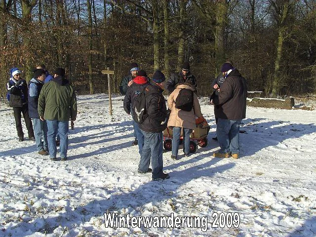 Gruppen von Menschen im Schnee, einige mit Hüten und Jacken, stehen in einem Waldgebiet, vermutlich bei einer Winterwanderung im Jahr 2009.