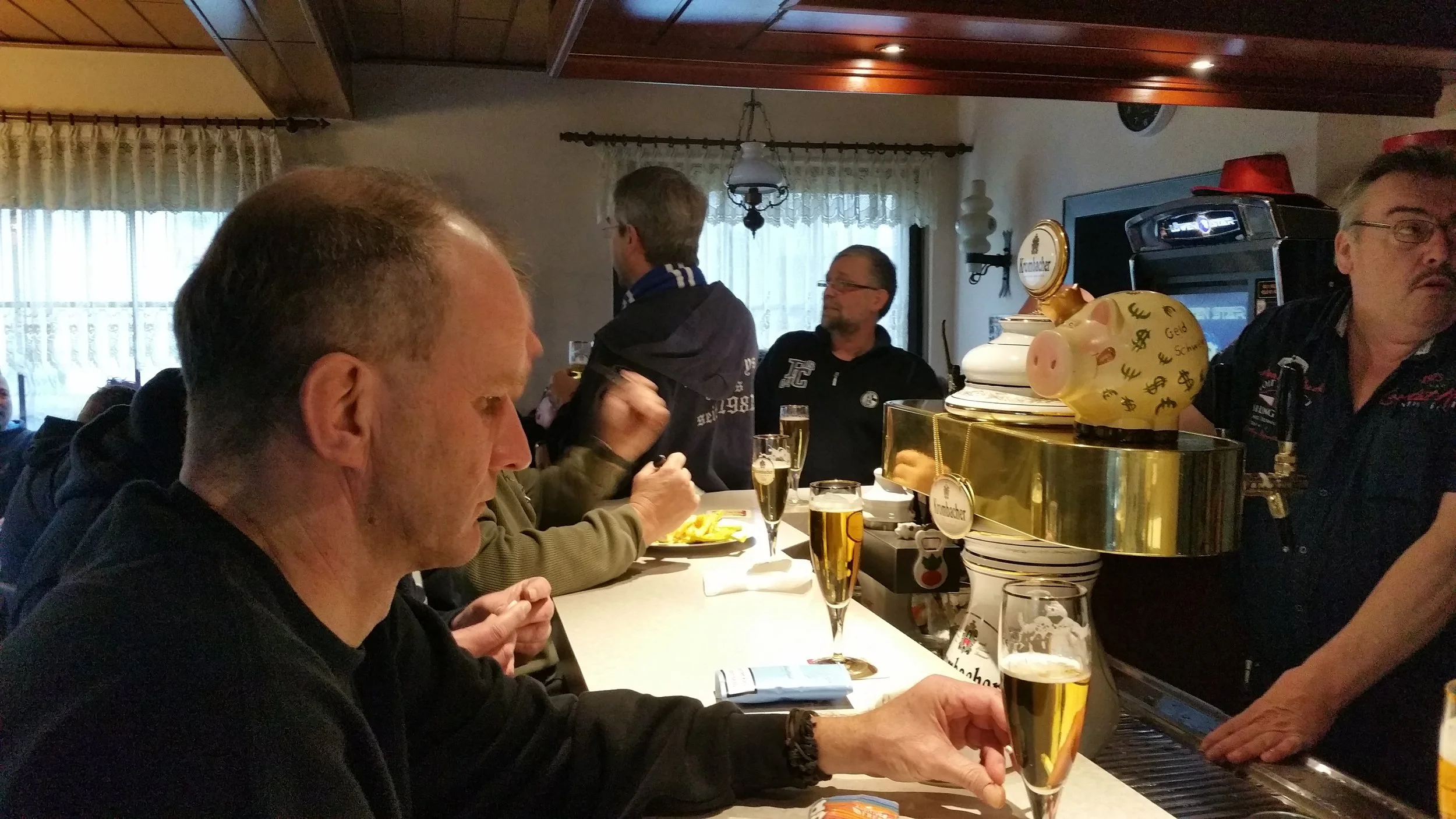 Menschen sitzen in einer gemütlichen Bar, trinken Bier und unterhalten sich. Es gibt eine dekorative Schweinefigur und ein Zapfhahn. Der Raum hat Fenster mit Spitzenvorhängen und Holzdecke.