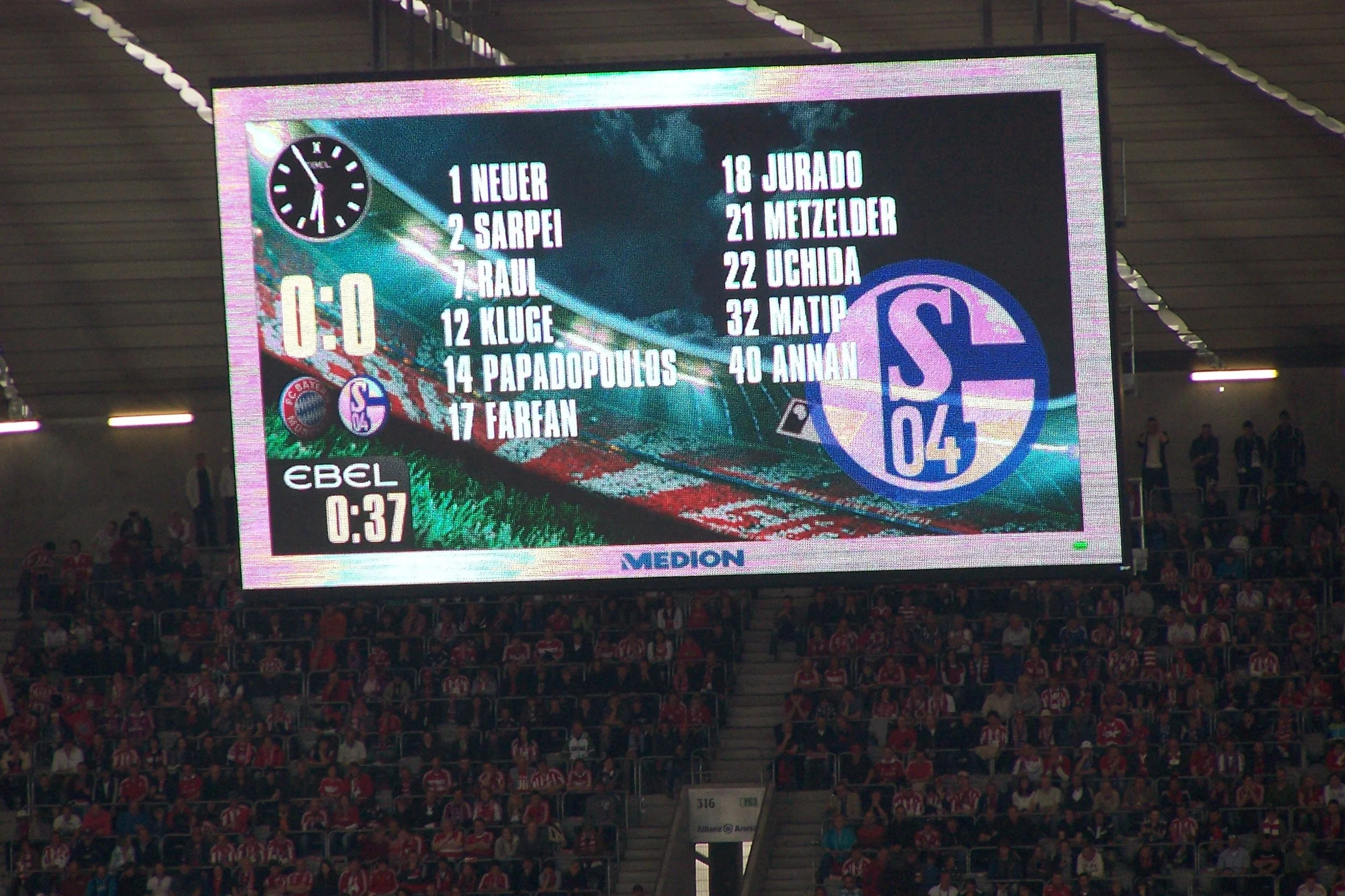 Ein Fußballstadion während eines Spiels, mit einer elektronischen Anzeigetafel, die das Ergebnis 1:0 anzeigt. Auf der Anzeigetafel sind die Namen der Spieler und ihre Nummern aufgelistet, mit einer Mannschaftskennung. Im Hintergrund sind viele Fans i