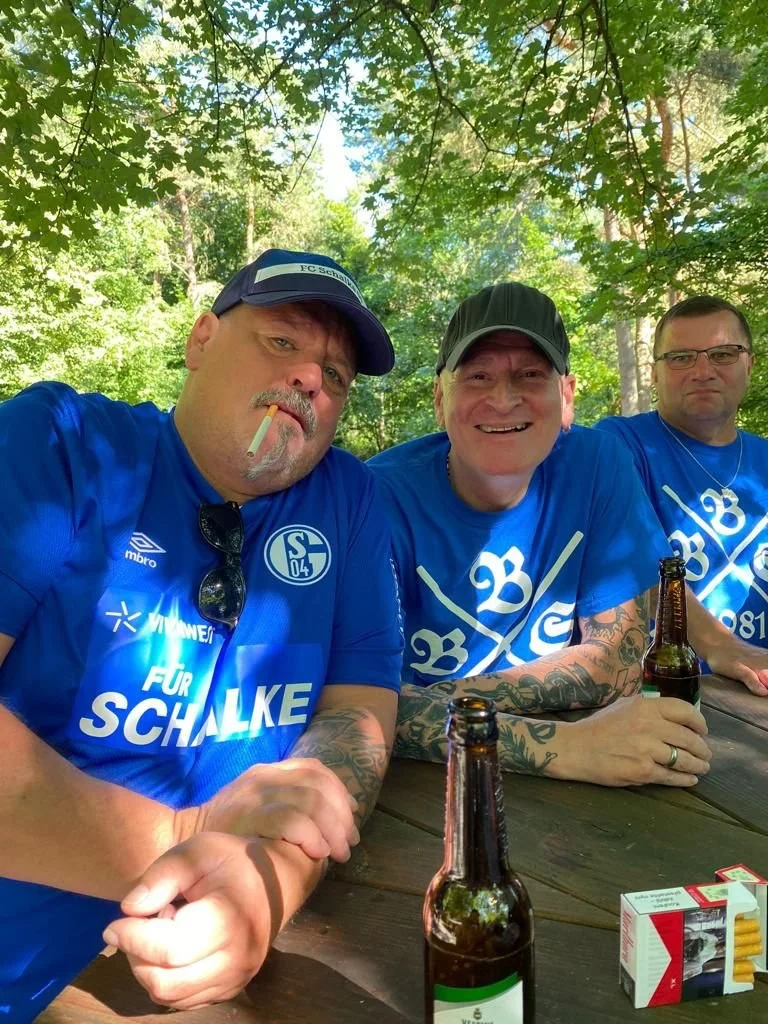 Drei Männer in blauen Schalke-Trikots sitzen an einem Holztisch im Freien, trinken Bier und genießen einen Tag in der Natur, umgeben von Bäumen und Sonnenlicht.