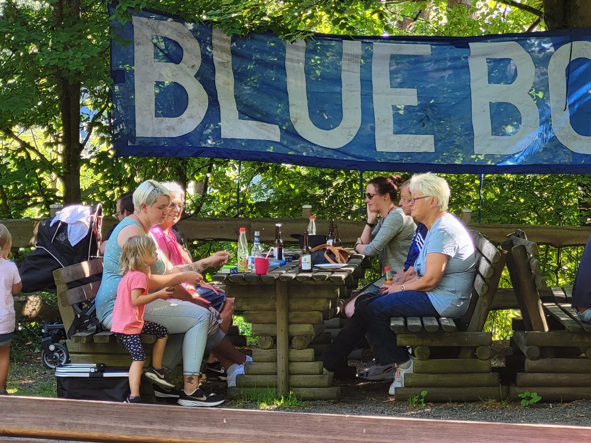 Menschen sitzen in einem Garten unter Bäumen an einem längeren Holztisch und genießen Getränke und Gespräche. Im Hintergrund hängt ein blauer Banner mit weißen Buchstaben.