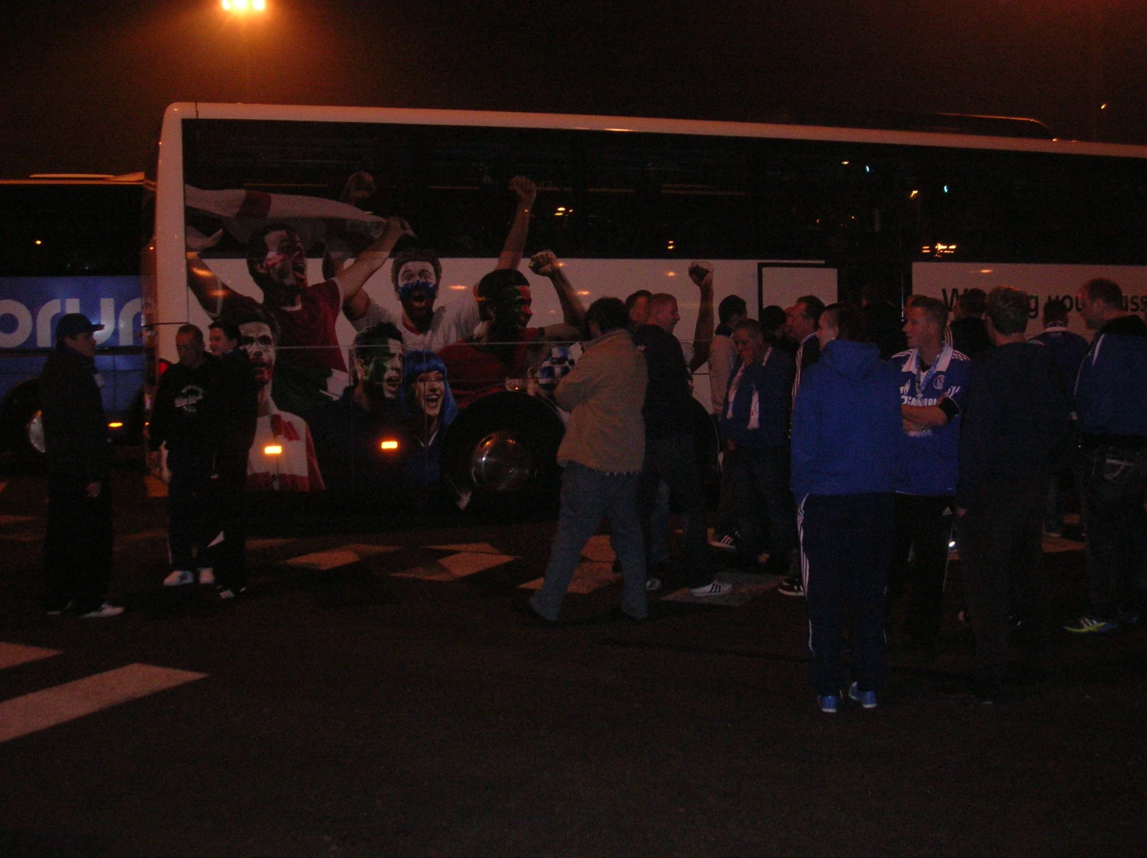 Menschenmenge vor einem Bus in der Nacht, einige Menschen jubeln und machen Handzeichen, im Hintergrund Dunkelheit und Straßenbeleuchtung.