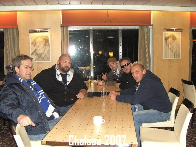 Fünf Männer sitzen an einem Holztisch in einem Restaurant, einige tragen Chelsea Football Club Schal. Im Hintergrund sind Fenster mit Vorhängen und Porträtbilder an der Wand.