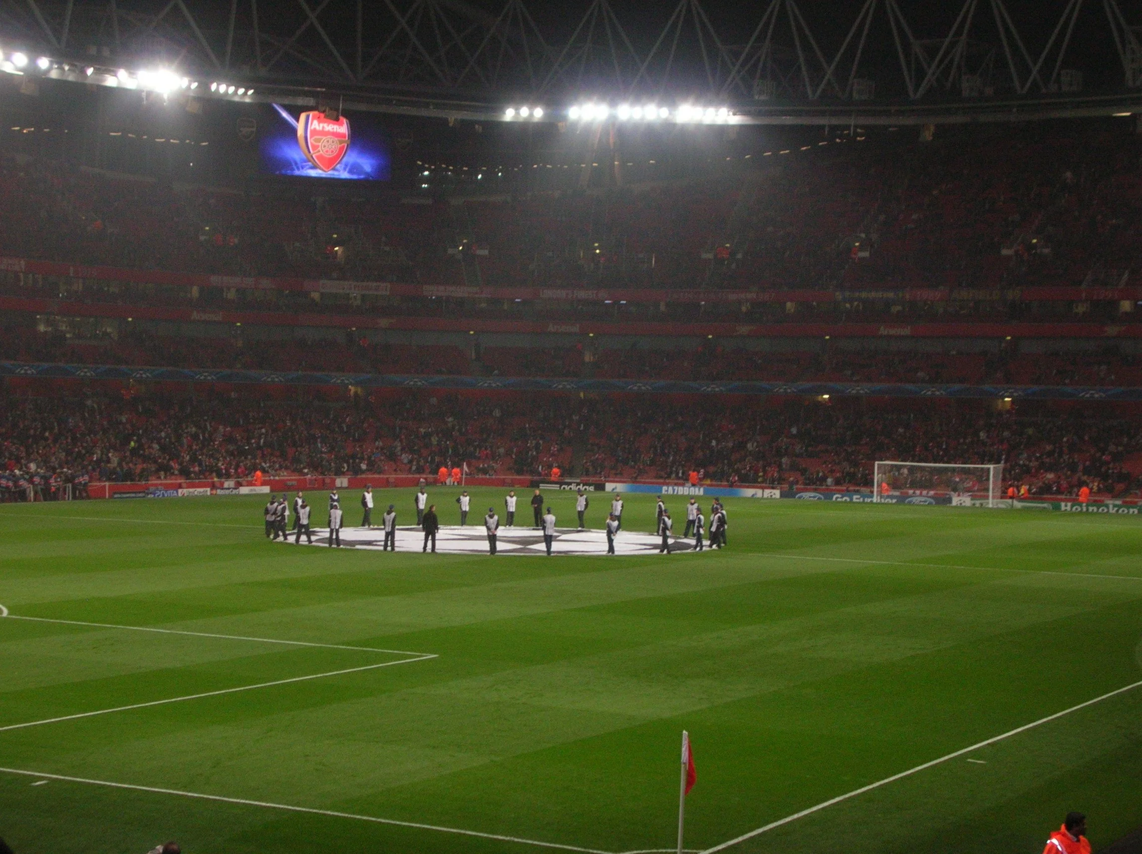 Ein Fußballstadion während eines Spiels mit der UEFA Champions League, es sind Spieler auf dem Feld, und im Hintergrund ist das Vereinslogo von Arsenal auf dem großen Bildschirm zu sehen.