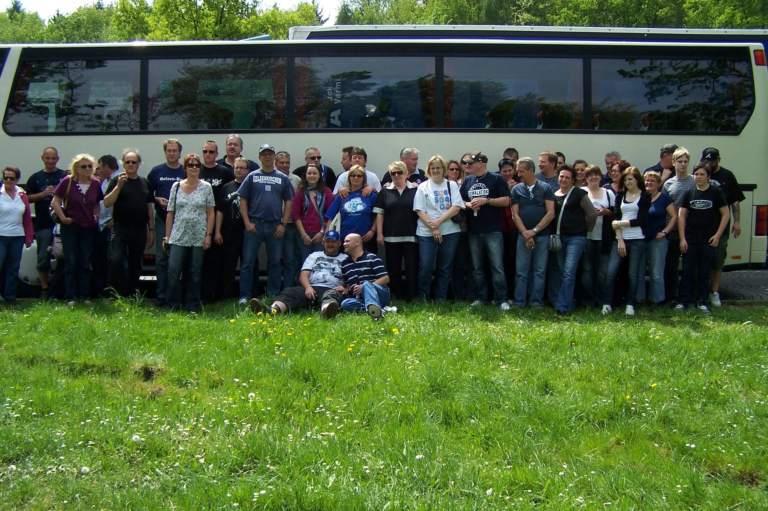 Gruppe von Menschen vor einem weißen Reisebus auf einer grünen Wiese an einem sonnigen Tag.