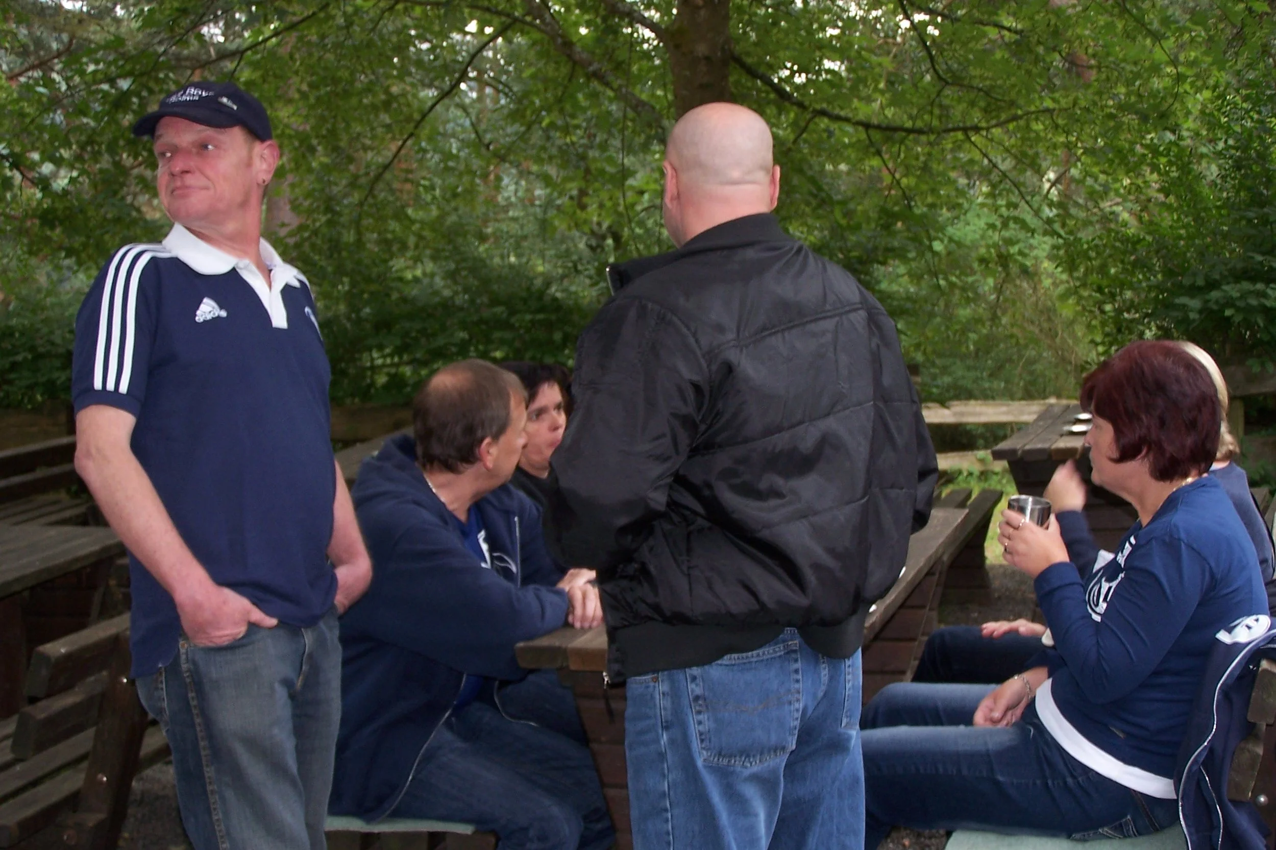 Menschen sitzen und sprechen in einem Park, einige stehen, umgeben von grünen Bäumen.