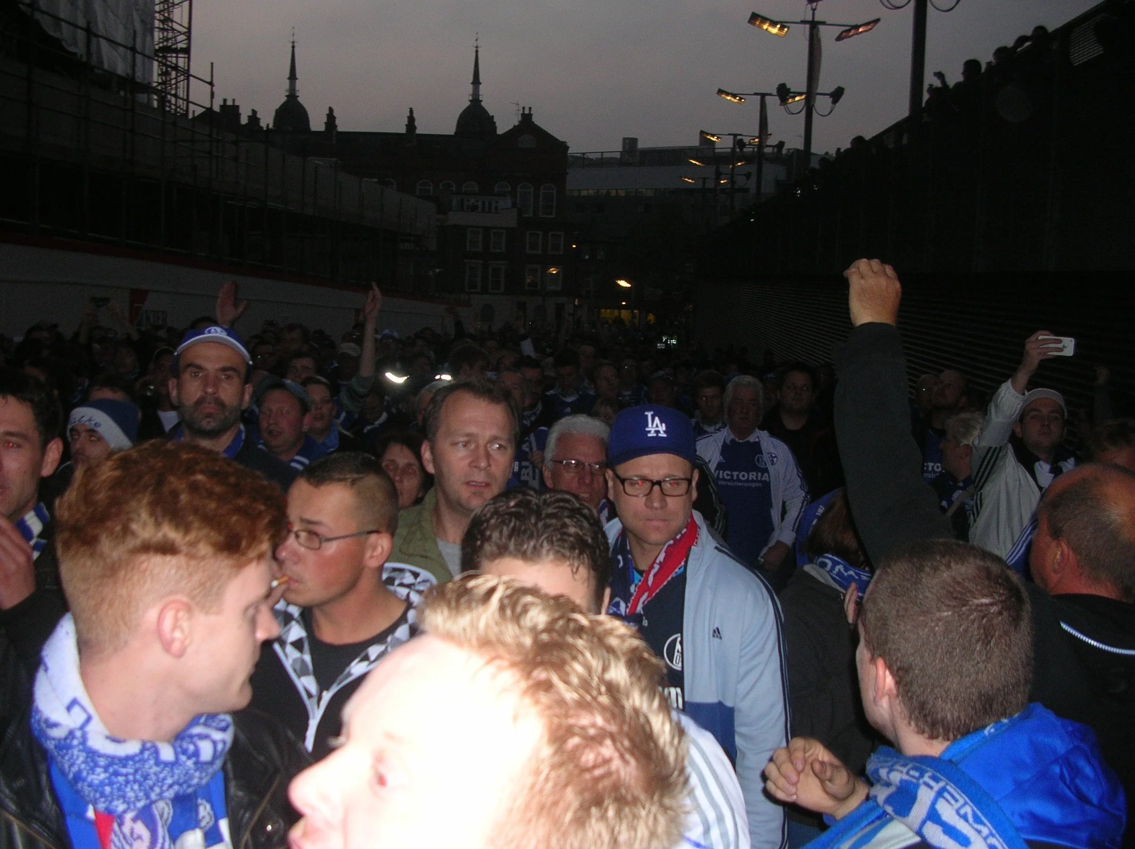 Menschenmenge beim Stadionbesuch, viele Fans tragen blaue Kleidung und Mützen, in einer dunklen Umgebung, wahrscheinlich bei Dämmerung oder Nacht.