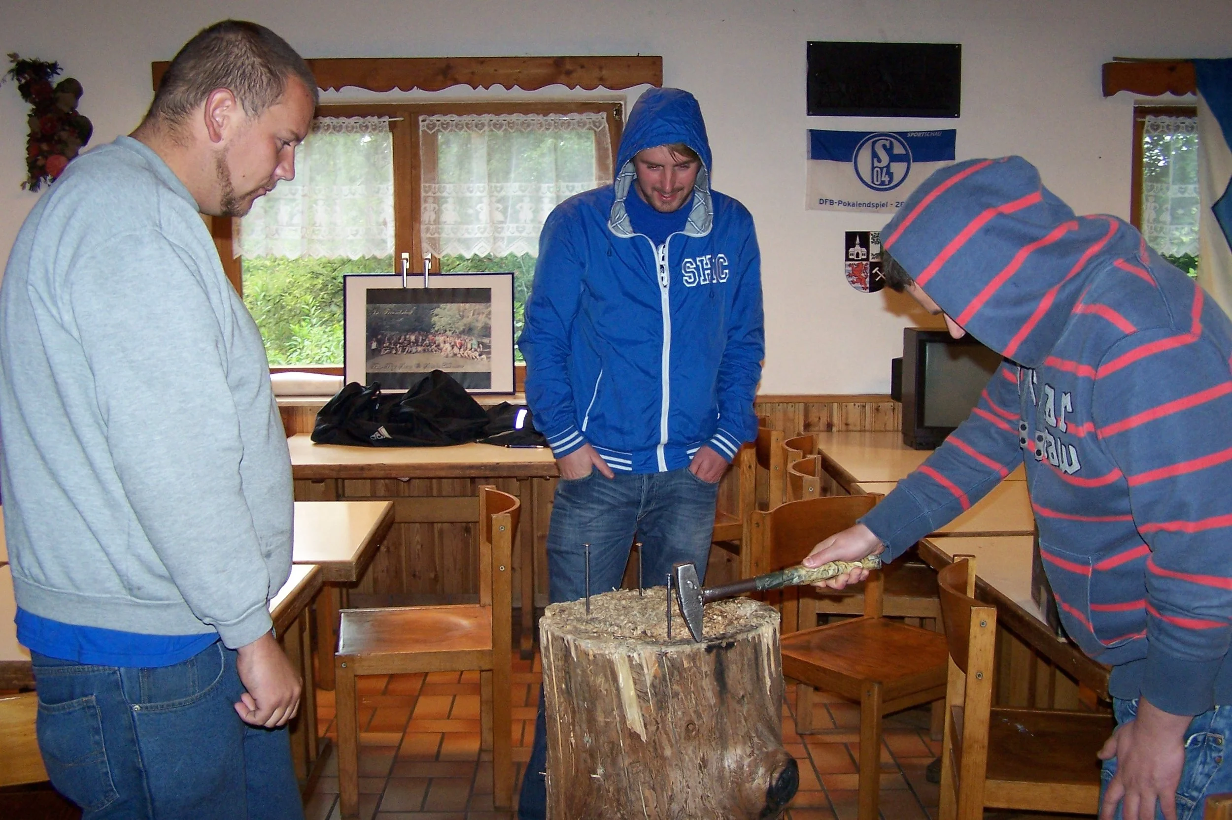 Drei junge Männer stehen um einen Baumstamm, einer schlägt mit einer Axt auf den Baumstamm, während die anderen beiden zuschauen. Es sieht nach einer Holzarbeit im Innenraum aus.