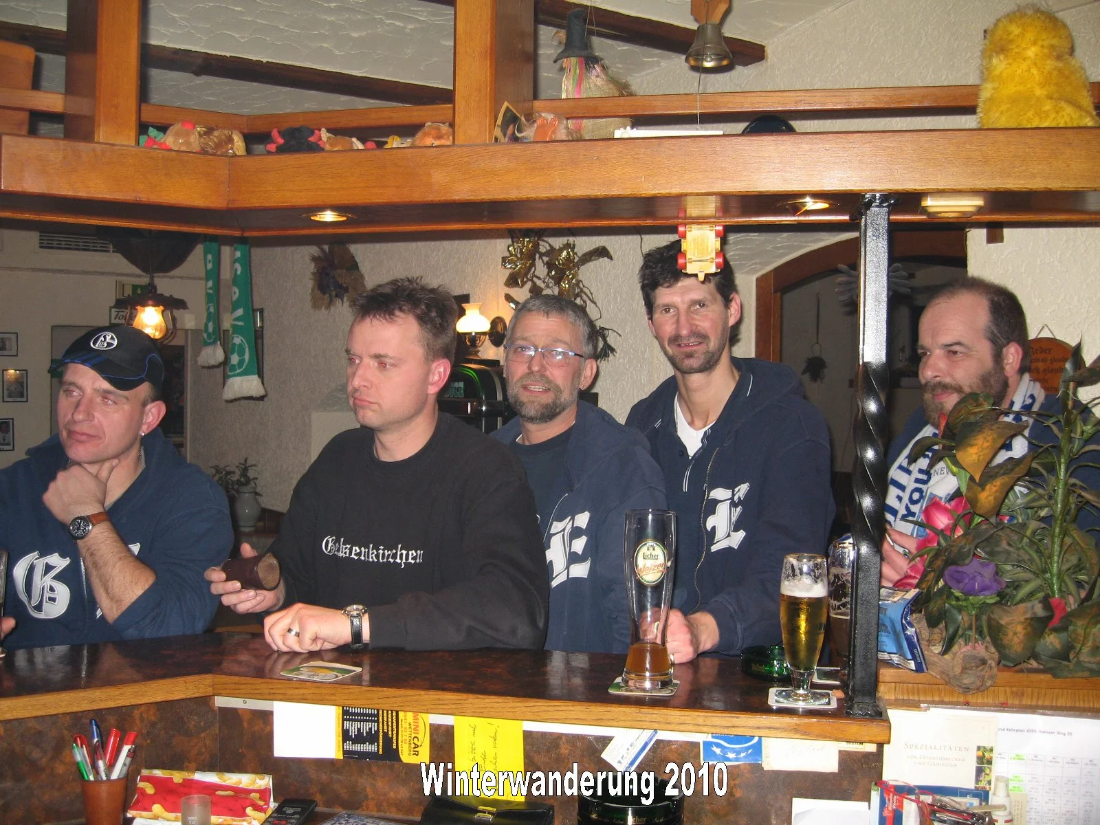 Fünf Männer sitzen an einem Stehtisch in einer rustikalen Bar. Sie haben Getränke vor sich, einige mit Bier. Der Hintergrund zeigt eine Holzdecke, dekorative Gegenstände und eine Blume. Der Text 'Winterwanderung 2010' ist am unteren Bildrand.