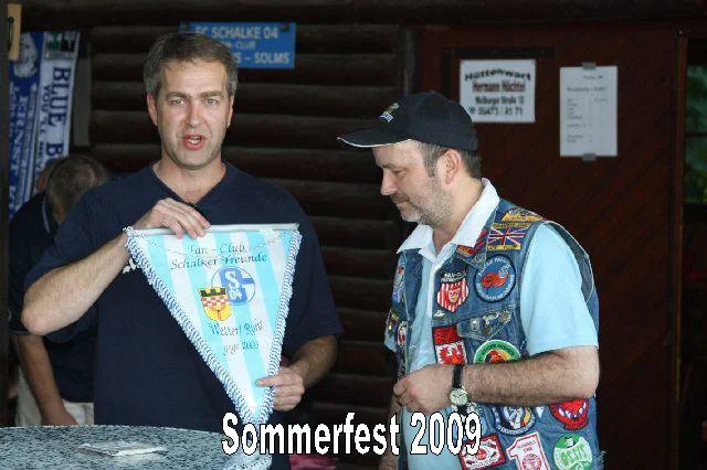 Zwei Männer sprechen bei Sommerfest 2009, einer hält ein blau-weißes Wimpelband. Der andere trägt eine Jean Jacke mit Abzeichen, die auf einem Holzgebäude im Hintergrund stehen.