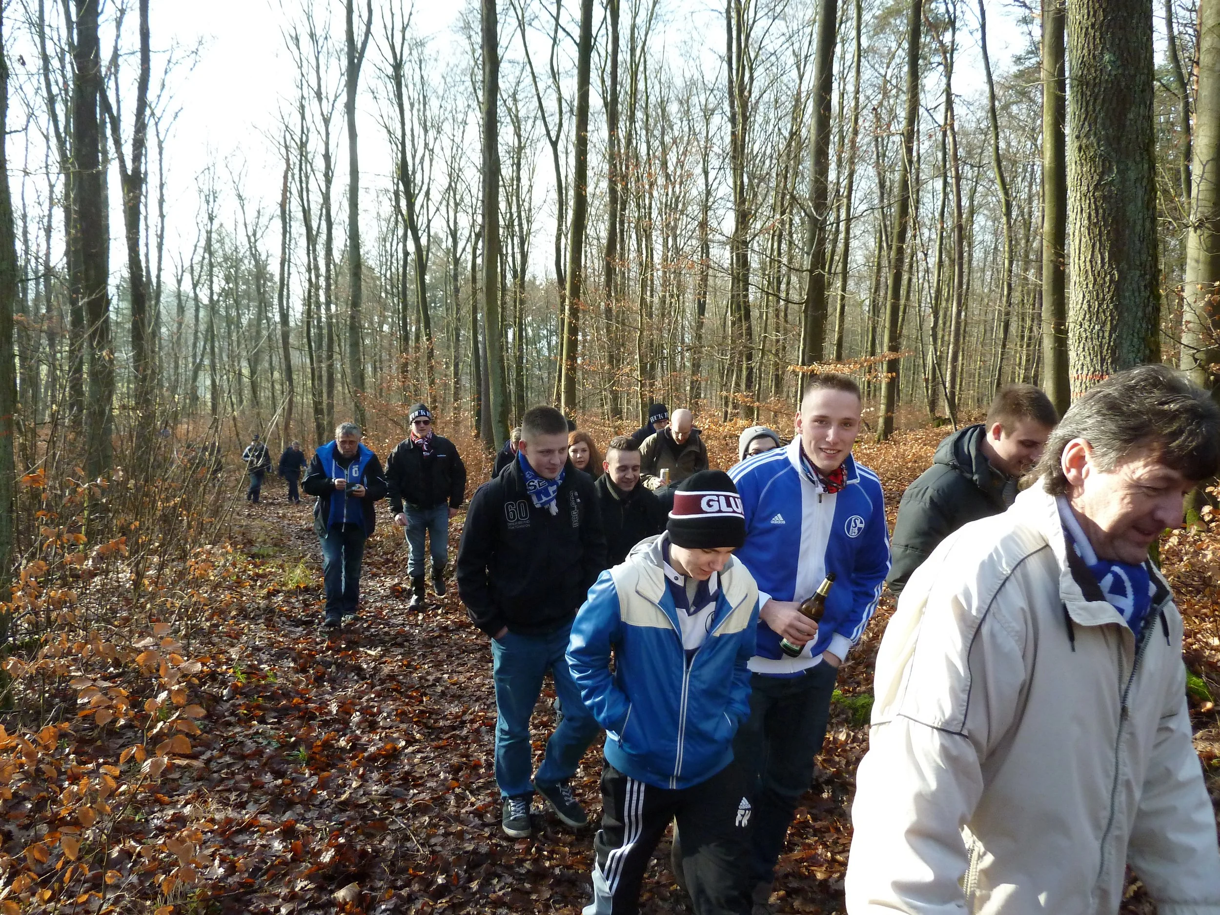 Eine Gruppe von jungen Menschen wandert durch einen herbstlichen Wald, viele tragen Sportswear und einige haben Getränke in der Hand.