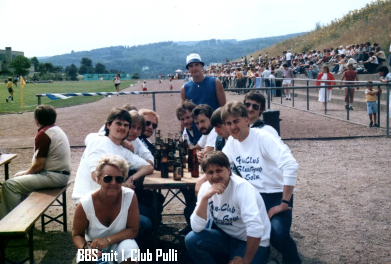 Gruppe von jungen Menschen und einer älteren Frau sitzen an einem Holztisch im Stadion auf der Tribüne, im Hintergrund ein Fußballfeld und Zuschauer. Es ist ein sonniger Tag.