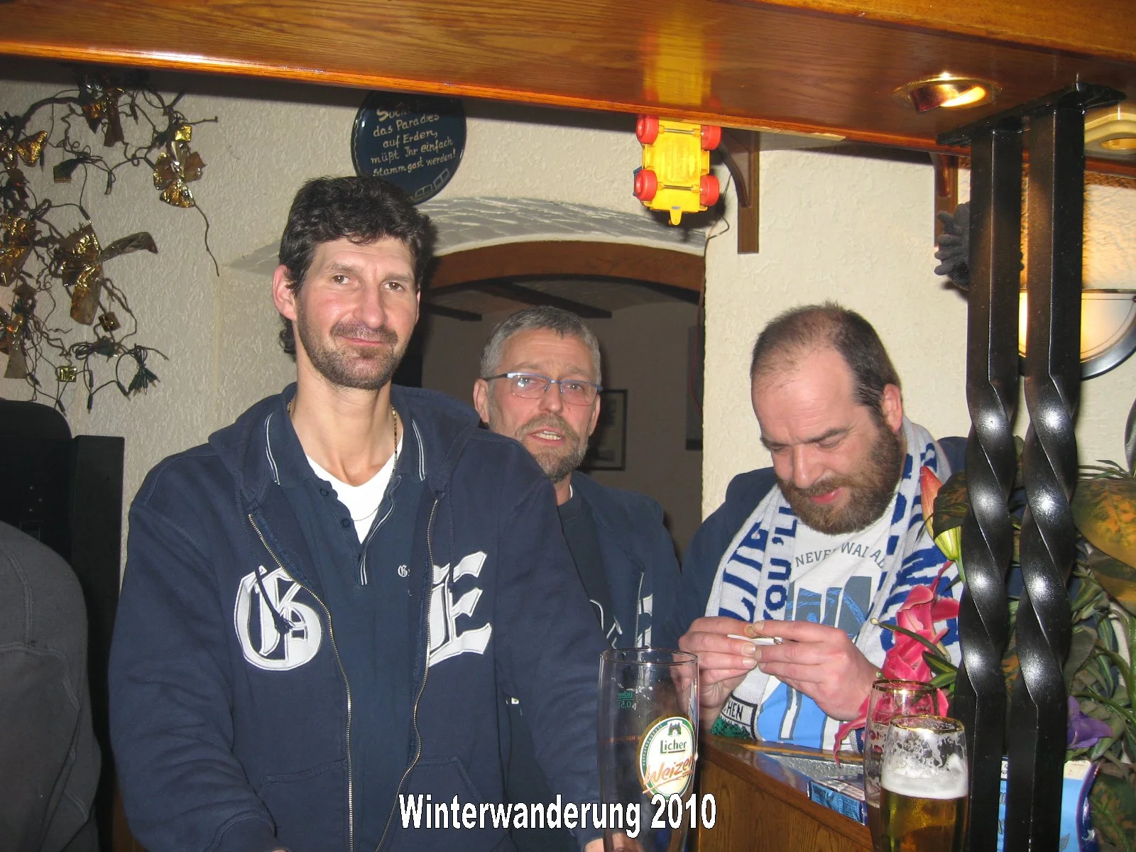 Drei Männer sitzen an einem Tresen in einer Bar, trinken Bier und unterhalten sich. Im Vordergrund steht ein Mann mit dunklen Haaren und Bart, im Hintergrund sind zwei Männer, einer mit Brille, der andere mit einem Bart und Trageohrring. Es befindet 