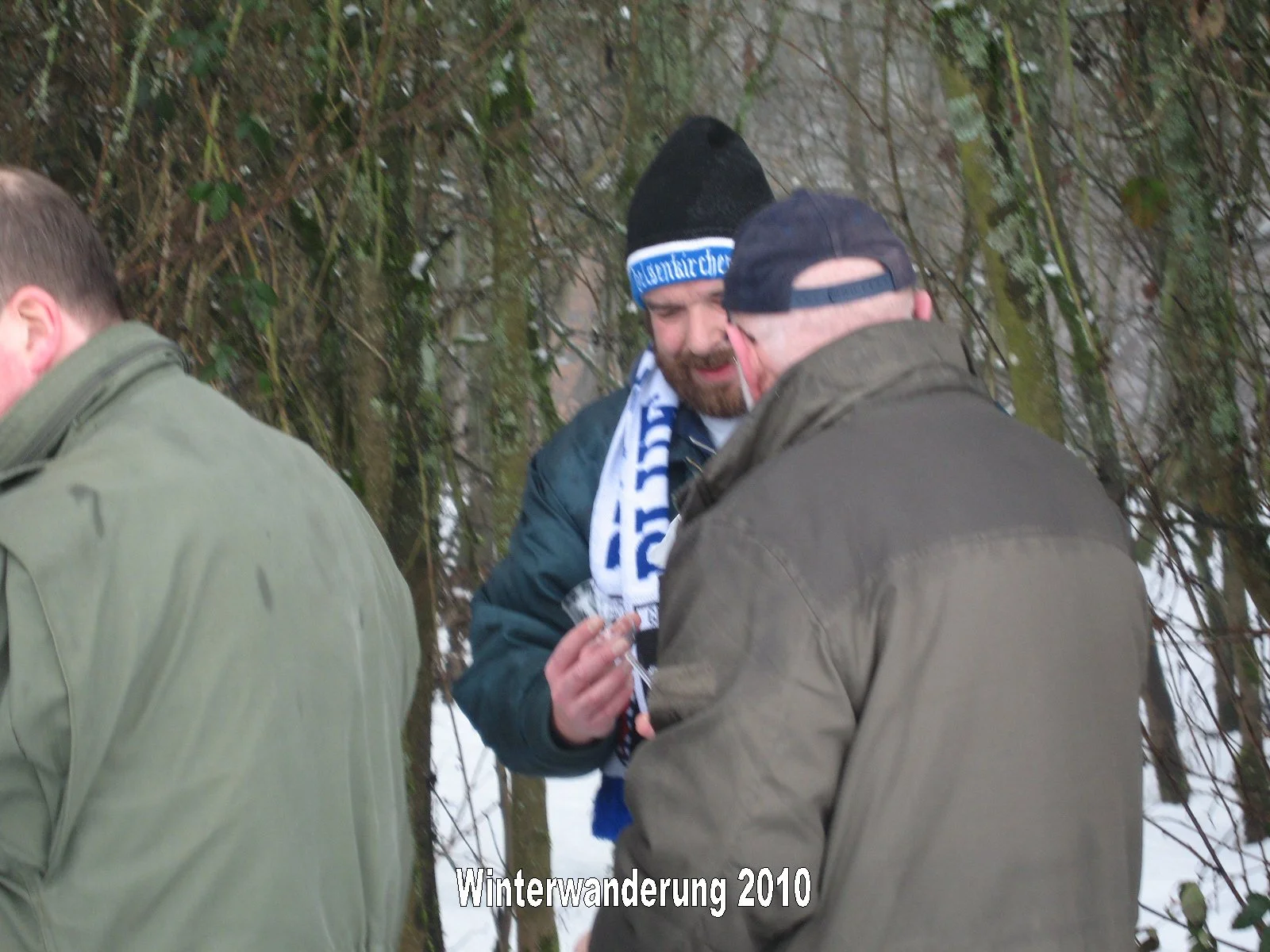 Drei Männer stehen im Schnee in einem Wald und unterhalten sich. Einer trägt eine schwarze Mütze mit blauer Aufschrift, ein anderer eine Mütze. Der Text "Winterwanderung 2010" ist am unteren Rand des Bildes zu sehen.